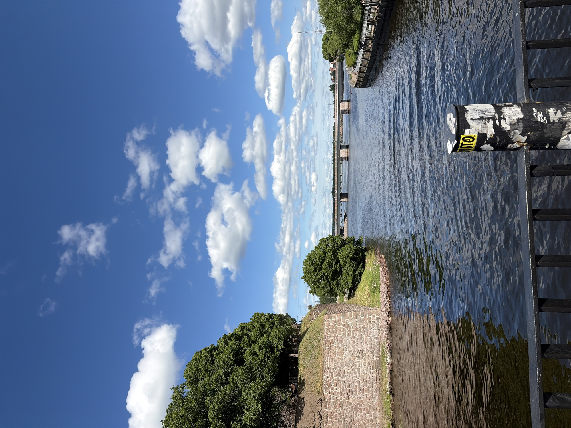 A wide shot captures a sunny day with a bright blue sky dotted with fluffy white clouds. The foreground is dominated by a black metal railing and a cylindrical bollard covered in peeling stickers, one of which prominently displays the word "JUSTO." To the left, a stone wall, part of a historical structure, rises from the water's edge and is topped by lush green trees. The water is a dark blue, reflecting the sky and the surrounding greenery. A long bridge with multiple arches stretches across the water in the mid-ground, connecting two sides of the city. On the right bank, a paved promenade with decorative railings is visible, lined with trees and a few distant figures. The overall scene suggests a peaceful urban landscape with historical elements.