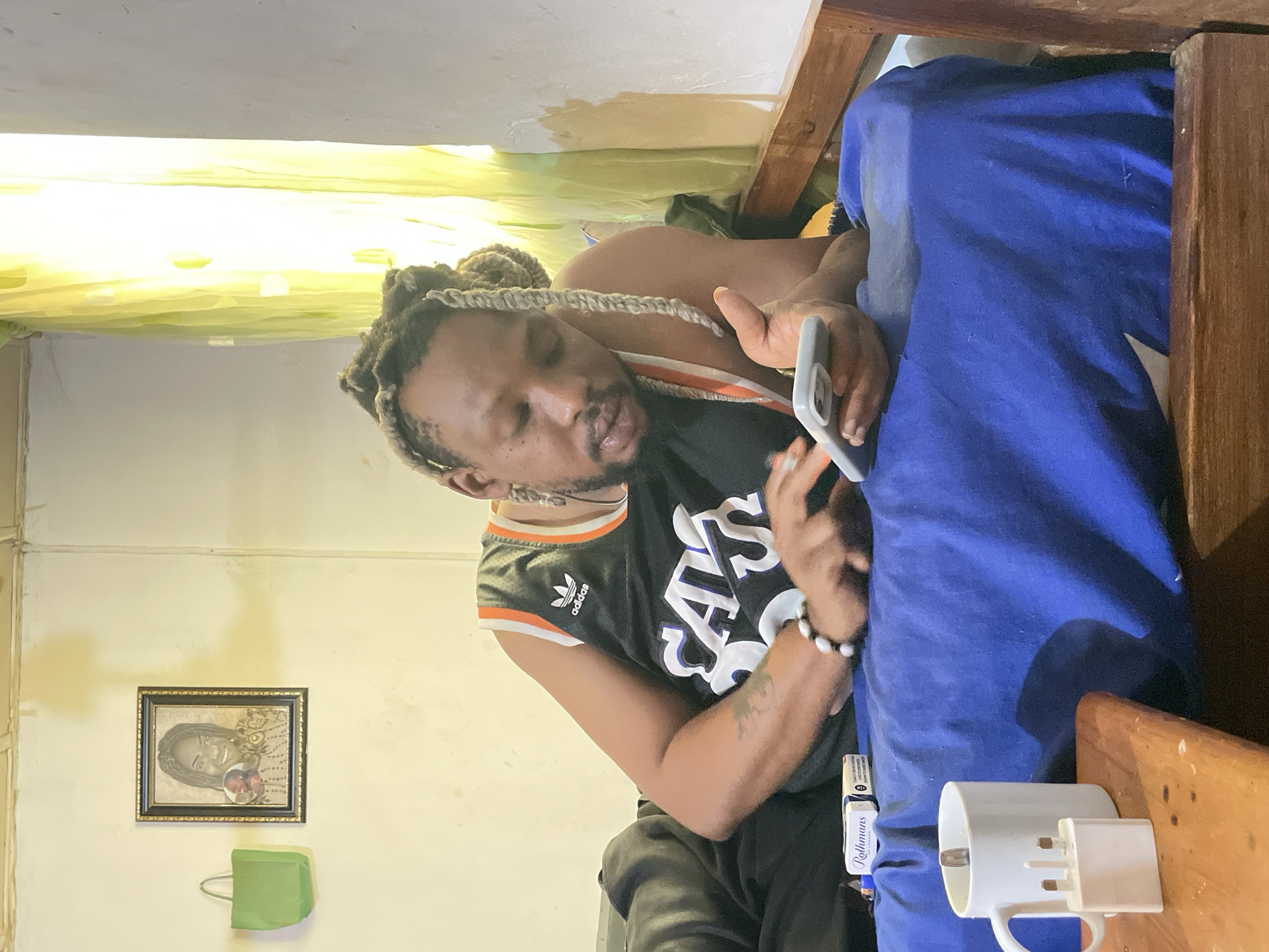 A man with dreadlocks is lying in bed, holding a smartphone. He is wearing a black Adidas jersey with white lettering and orange trim. A beaded bracelet adorns his left wrist. He appears to be looking at or interacting with the phone.
The bed has blue bedding. A white mug containing an electrical adapter is placed on the bed near the man. A pack of Rothmans cigarettes rests near the mug.
The room has a faded yellow-white wall. On the wall, there is a framed portrait of a woman and a green bag hanging below it.
The location is likely in Jos, Nigeria.