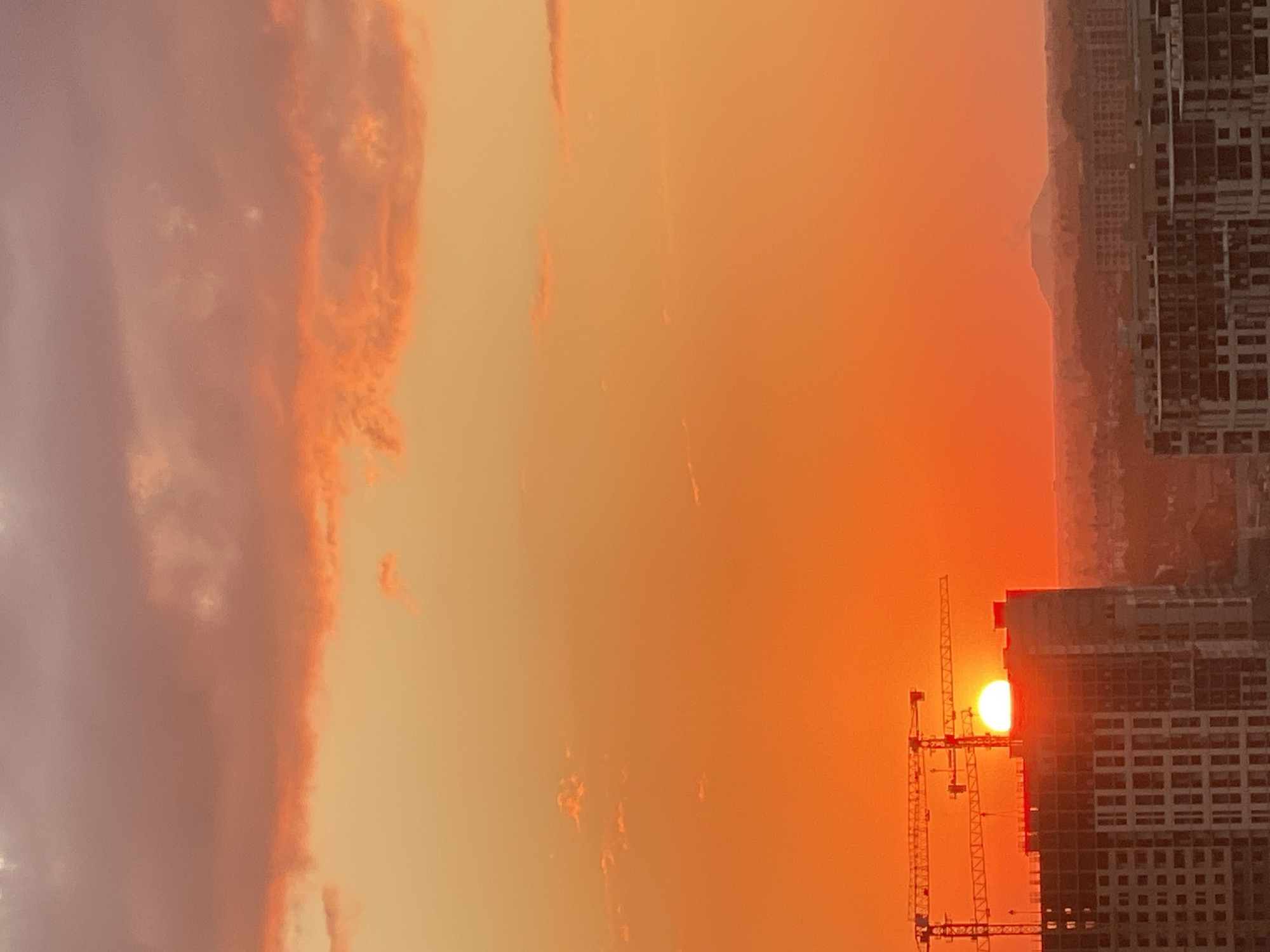 An extreme wide shot captures a vibrant sunset over a city. The sky is awash in fiery oranges and soft pinks, with scattered clouds catching the last rays of sunlight. In the foreground, the skeletal structures of a high-rise building under construction dominate the lower left, with cranes silhouetted against the bright sky. Further in the distance, the urban landscape stretches out, with rows of buildings and a lone, distant mountain visible on the horizon. The scene is devoid of any people.