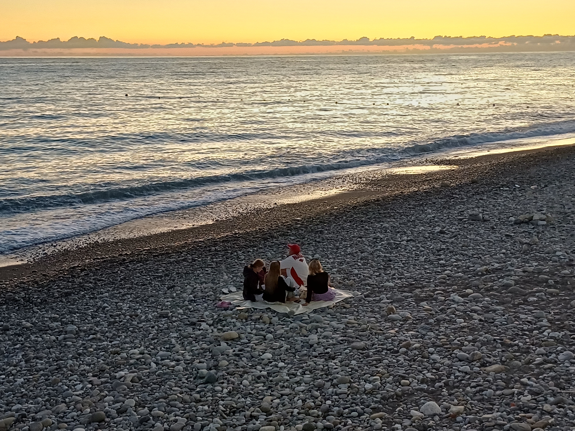 At sunset on a pebble beach in Sochi, Russia, a group of four individuals is seated on a blanket. One person, appearing to be a male wearing a red cap and a white shirt with red accents, is in the center, facing away from the camera. Three other individuals, two women and a child, are positioned around him, also facing inward. The sea is calm, with gentle waves lapping at the shore, and the sky is illuminated with hues of orange and yellow from the setting sun, with a line of clouds in the distance. The pebble beach is covered in smooth, gray stones of various sizes.