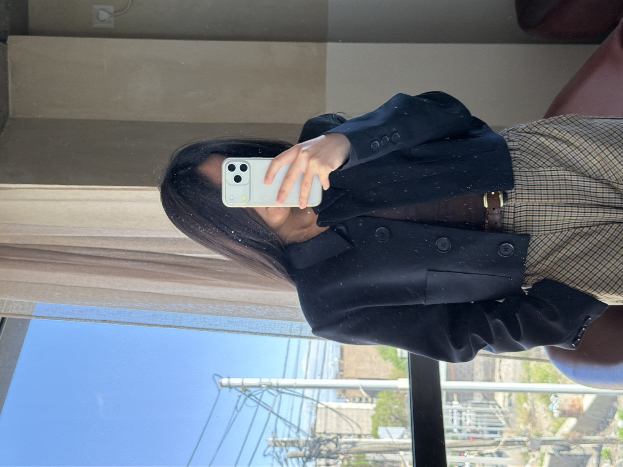 A person is holding a smartphone, likely taking a selfie, with a black blazer over a plaid skirt. A belt is visible at the waist. The reflection in the window shows a city scene with power lines, buildings, and greenery, possibly in Yerevan, Armenia. The window is partially covered by curtains. The view outside suggests an urban residential area.