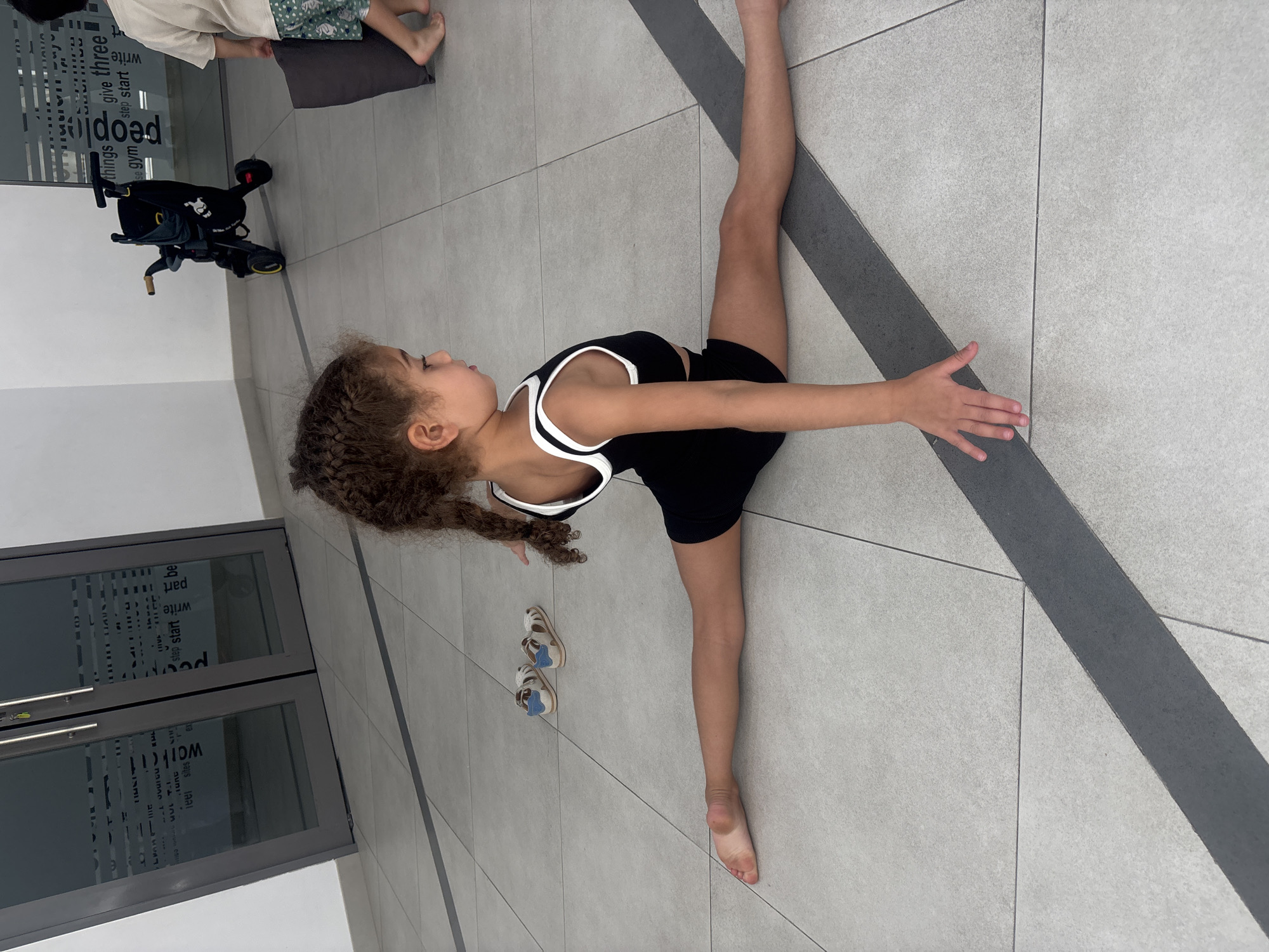 This media file shows a young girl performing a gymnastic split on a tiled floor. She is wearing a black and white athletic outfit. Her arms are extended outwards, and her legs are stretched in opposite directions, demonstrating impressive flexibility. A pair of sandals lies on the floor near her. In the background, a stroller is visible, along with a glass door with text decals. The text on the door includes phrases like "give three," "step start," "write," and "part." The overall setting appears to be an indoor space, possibly a studio or a common area, with light grey tiled flooring and a dark grey diagonal stripe. The lighting suggests it is daytime. The girl's expression is focused and determined, indicating concentration on her gymnastic pose. The scene is set in Dubai, United Arab Emirates.