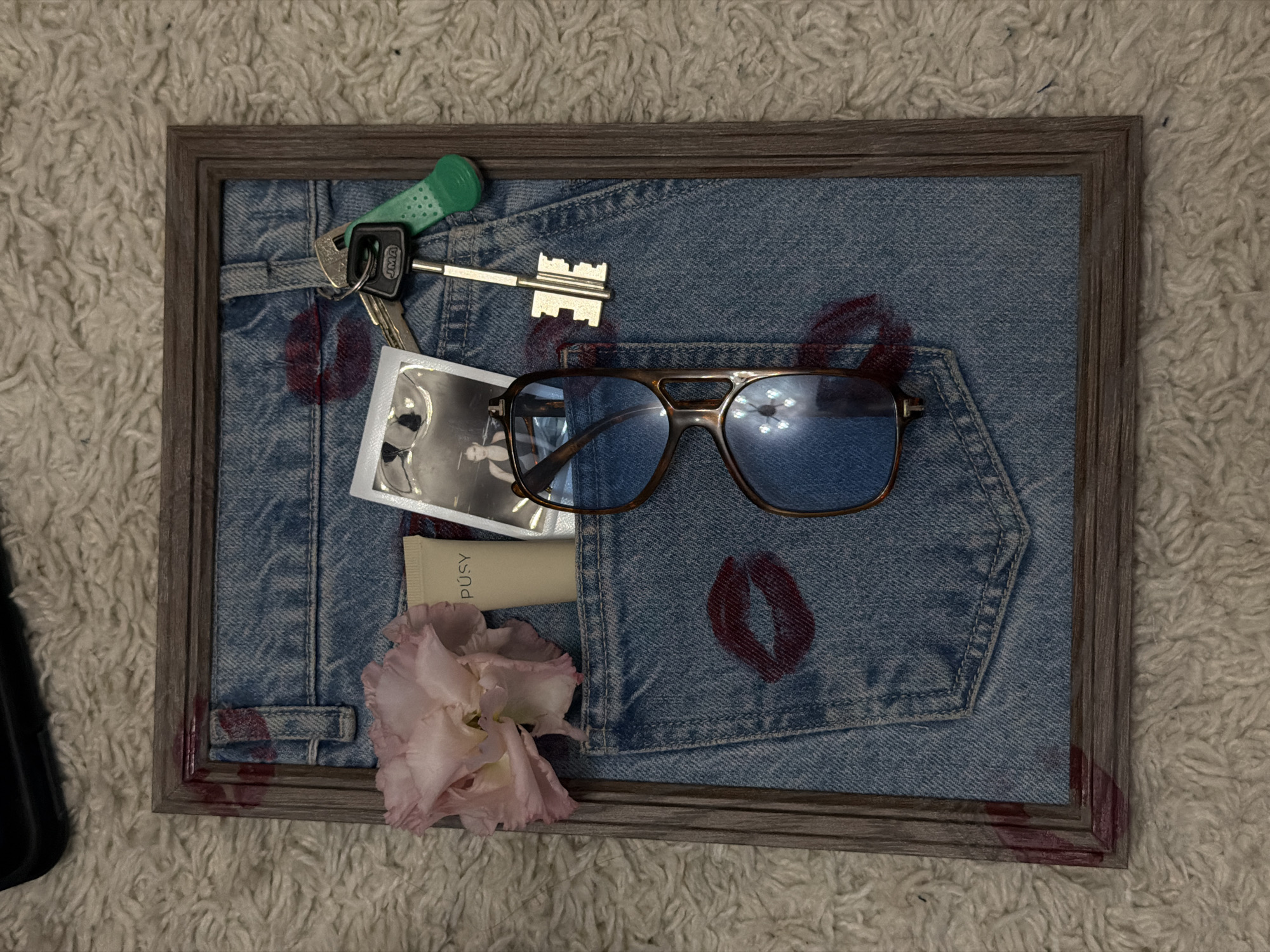 A framed arrangement rests on a shaggy, off-white surface. The frame is a weathered gray wood. Within the frame is a section of denim, likely from jeans, marked with multiple red lipstick kiss prints. A pair of aviator-style sunglasses with brown frames and blue lenses sits in the center of the denim. A key ring with several keys, one with a green handle and another resembling a castle key, is near the top left. A small, unlabeled tube of cream-colored lotion is tucked into a pocket on the denim. A pale pink flower is positioned in the lower left corner. There is also a small photograph in the scene. The photograph is likely a snapshot of a person. A black object sits partially visible in the lower left outside of the frame. The scene does not obviously depict or suggest any association to Krasnoyarsk, Russia.