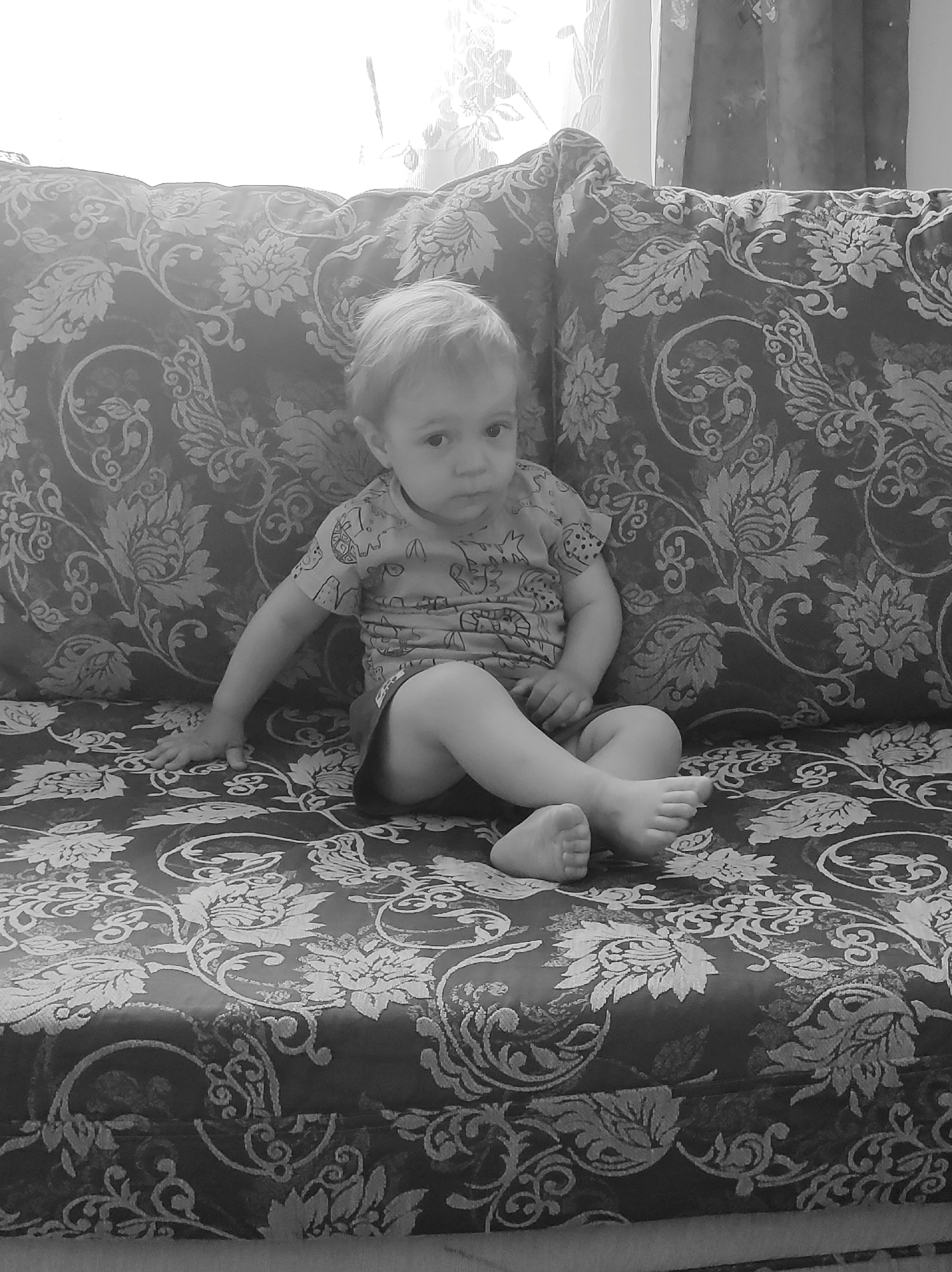 A black and white, eye-level medium shot features a baby sitting on a couch. The baby has fair skin and short, light-colored hair. The baby is wearing a shirt and shorts with patterns. The baby is sitting with legs crossed. The background features the couch and curtains. The couch has a dark floral pattern. The curtains are partially visible on the right side. The location is in a residential setting in the city of Unknown, Unknown.