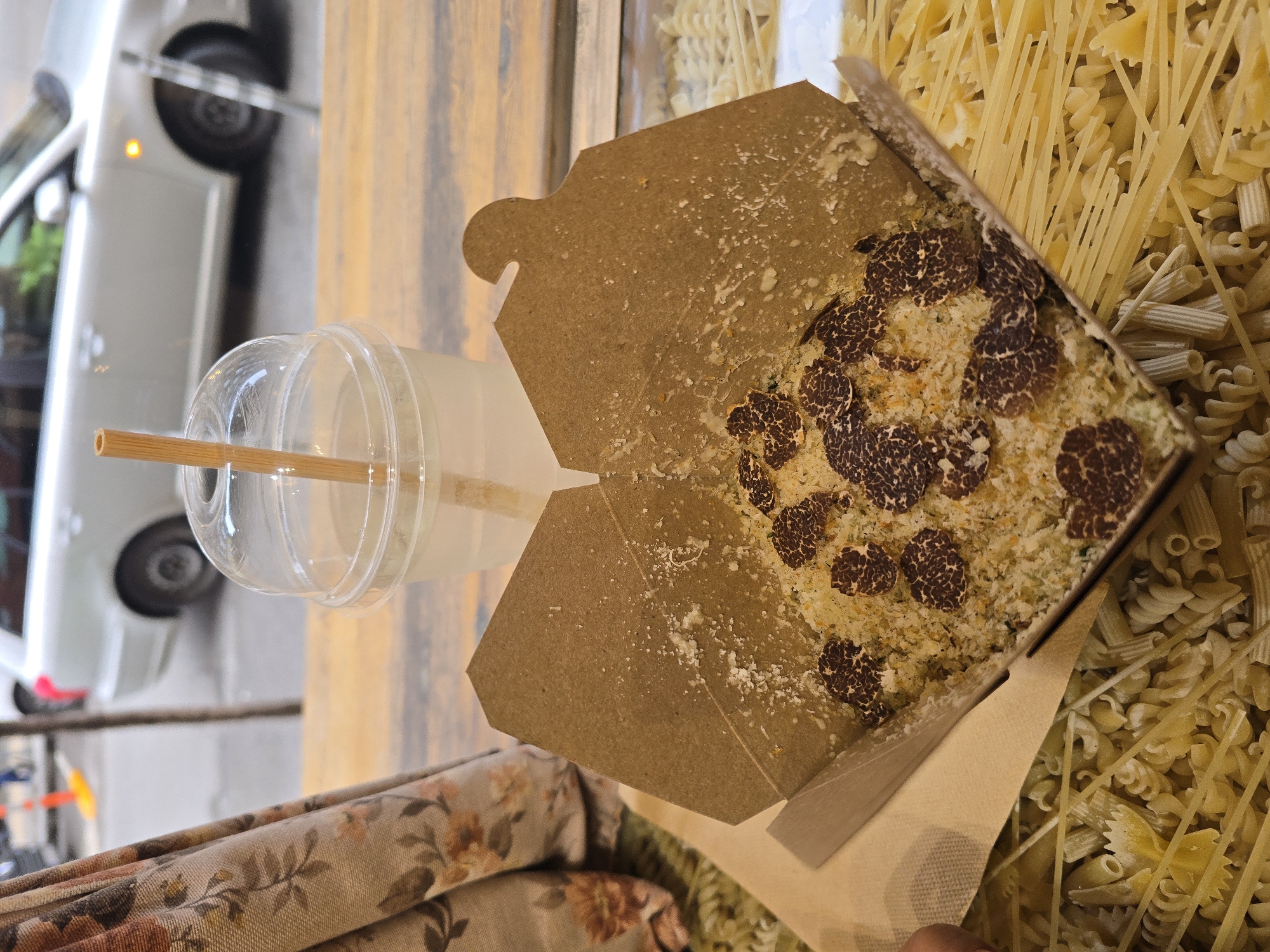 The image depicts a meal served in an open, light-brown corrugated cardboard takeaway container, containing a dish of small white pasta or rice, garnished with grated white cheese and multiple irregular, dark brown slices of black truffle. White granular residue adheres to the container's interior flaps. Adjacent to it, a clear plastic cup with a dome lid and a light-colored straw holds a translucent pale liquid. These items are positioned on a horizontally-grained wooden surface. Below this surface, a variety of uncooked pasta—spaghetti, fusilli, farfalle, and rigatoni—is scattered across the lower frame. A floral-patterned curtain, exhibiting brown, orange, and white hues, occupies the left margin. The scene is set within an establishment in Sofia, Bulgaria, looking out onto a blurred urban street. A light-colored sedan or station wagon is partially visible in the background, displaying its rear section and an illuminated amber indicator light. A slender vertical pole and an orange traffic cone are also present on the street. No individuals are clearly identifiable within the frame, and the specific street address in Sofia is not discernable from visual cues.