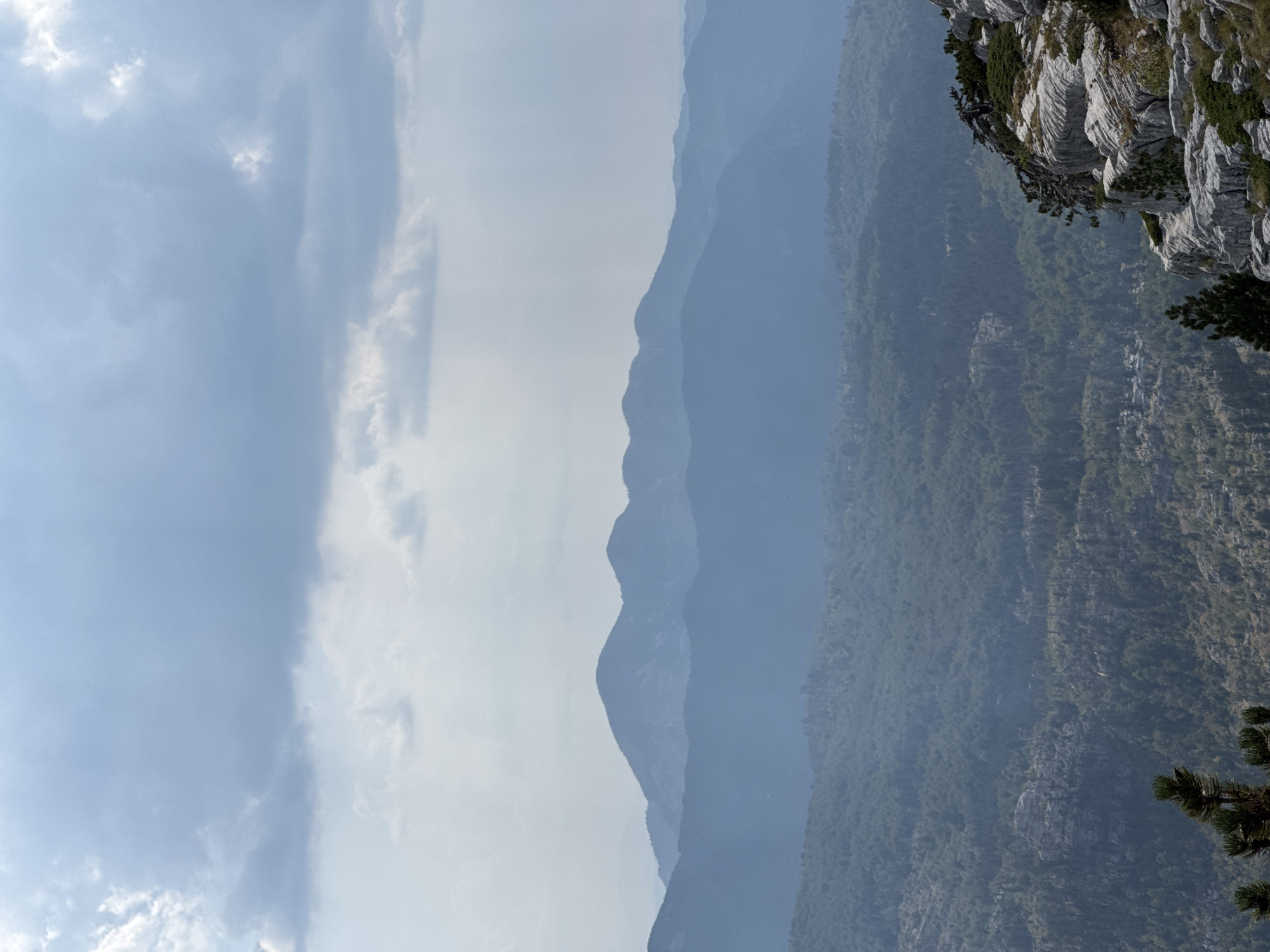 The image depicts a mountainous landscape under a cloudy sky. In the foreground, a rocky cliff face with sparse vegetation juts out into the frame. The middle ground is dominated by a densely forested mountainside, with trees covering the slopes. In the background, several layers of receding mountains are visible, gradually fading into a hazy blue. The sky is filled with a mix of light and dark clouds, suggesting an overcast or potentially stormy day. The scene appears to be an outdoor natural environment, likely a mountain range. There are no people or specific man-made structures clearly visible in the frame.