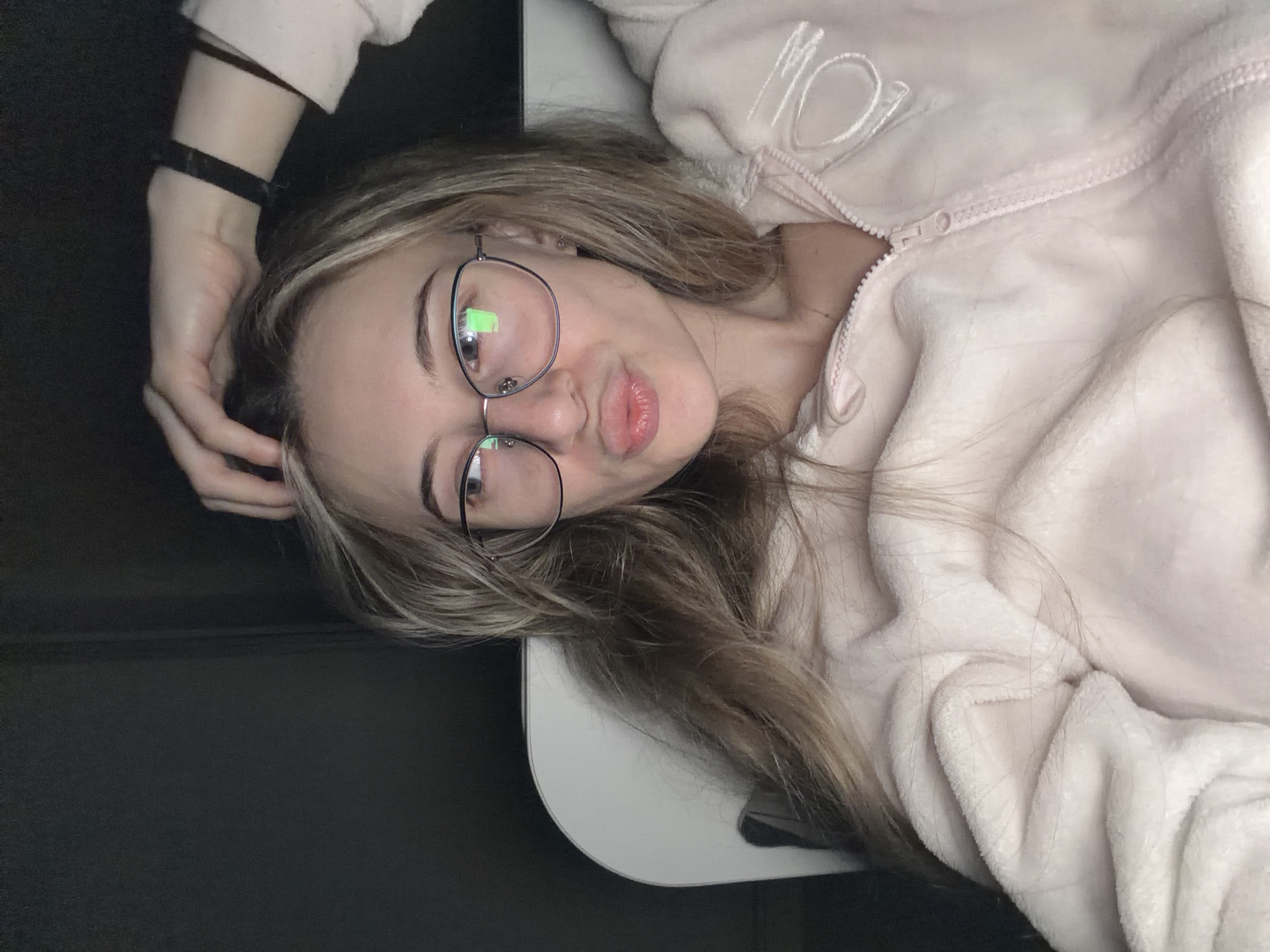 A young woman lies on a white surface. She wears glasses and a light pink zipped fleece jacket, and rests her left hand on her head. The location is not discernible, and there is no indication the image was taken in Plovdiv, Bulgaria. The background is dark and ambiguous.