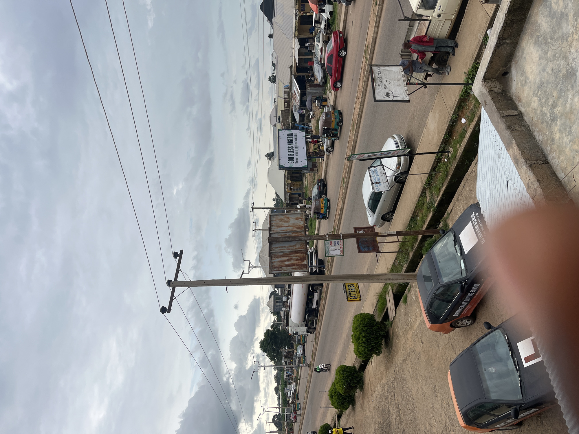 The image shows a street scene in Rafin Par, Nigeria. A road with multiple lanes is visible, with various vehicles including cars, a tanker truck, a motorcycle, and auto-rickshaws. Several people are present, some walking, some standing, and some driving. There are buildings lining the street, some with shops or businesses, and a large billboard with the text "GOD BLESS NIGERIA." Telephone poles with wires are also prominent. The sky is overcast, suggesting cloudy weather. The overall impression is that of a busy, everyday street in an urban or semi-urban area.