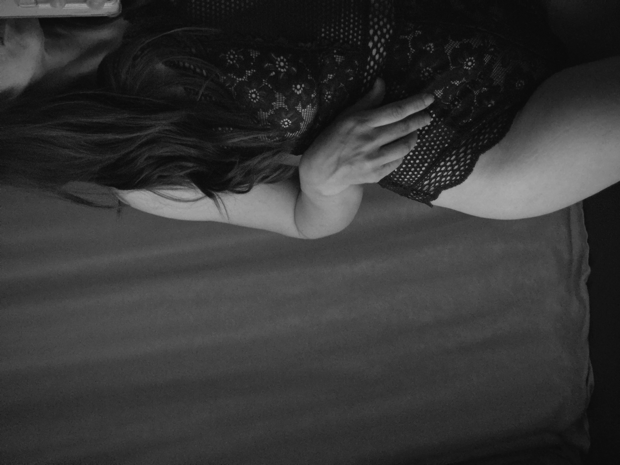 This monochrome image, captured from a top-down perspective, features a person lying down indoors, likely on a bed or a soft surface. The subject is wearing a dark, intricately patterned lace garment, resembling a bodysuit or lingerie, which features floral or geometric designs.
Long, dark hair is fanned out around the head and shoulder area, partially obscuring the face, though parts of the jawline and ear are visible at the upper left. An arm is bent, and a hand rests gently on the person's torso, near the hip, drawing attention to the texture of the lace and the subtle curves of the body. The skin of the arm and hip is softly illuminated, contrasting with the deeper tones of the garment and the dark background.
The setting appears to be a bedroom, with the person positioned on a plain, dark grey or black sheet or blanket. The lighting is soft and subdued, creating a sense of intimacy and tranquility. The black and white tonality enhances the focus on form, texture, and light, contributing to a calm and introspective mood. There is no visible text present in the image.