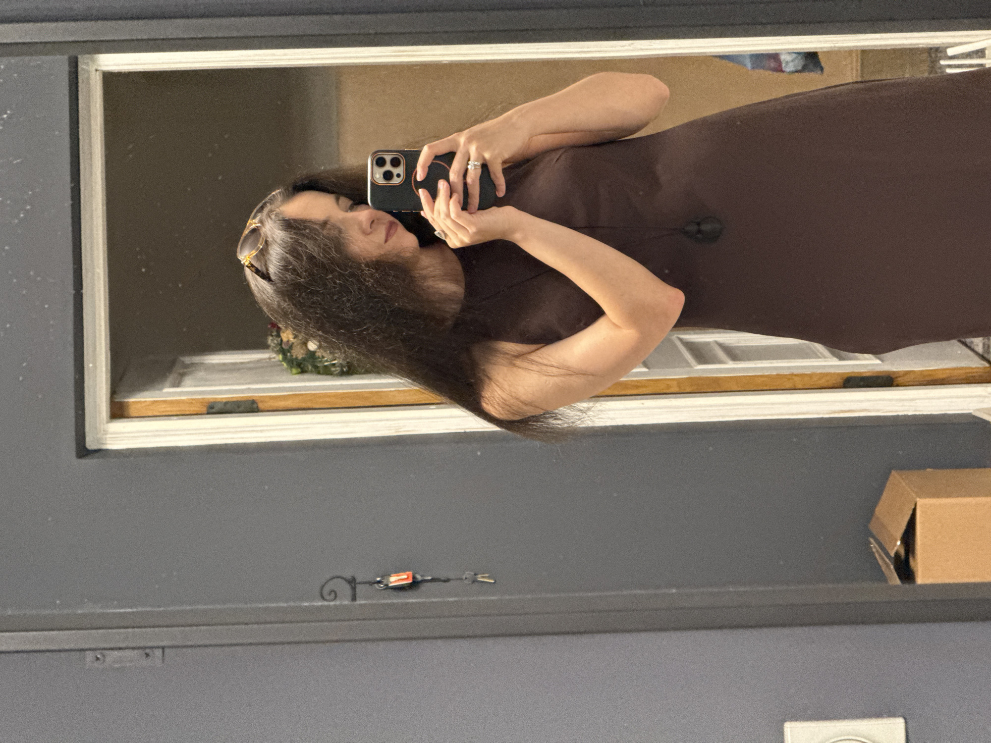 A young woman with long, dark, wavy hair and sunglasses perched on her head is taking a mirror selfie. She is wearing a sleeveless, dark brown dress and has a black pendant necklace. She is also wearing two rings on her left hand. In the background, a door frame with a white trim is visible, and a reflection shows another doorway with a glimpse of what appears to be a decorated wreath. To the left of the woman, a dark gray wall has a decorative metal hook with keys hanging from it. Below the hook, a small cardboard box is visible. The scene appears to be indoors, possibly in a home. The location is not in the city of Chaikovsky, Russia.