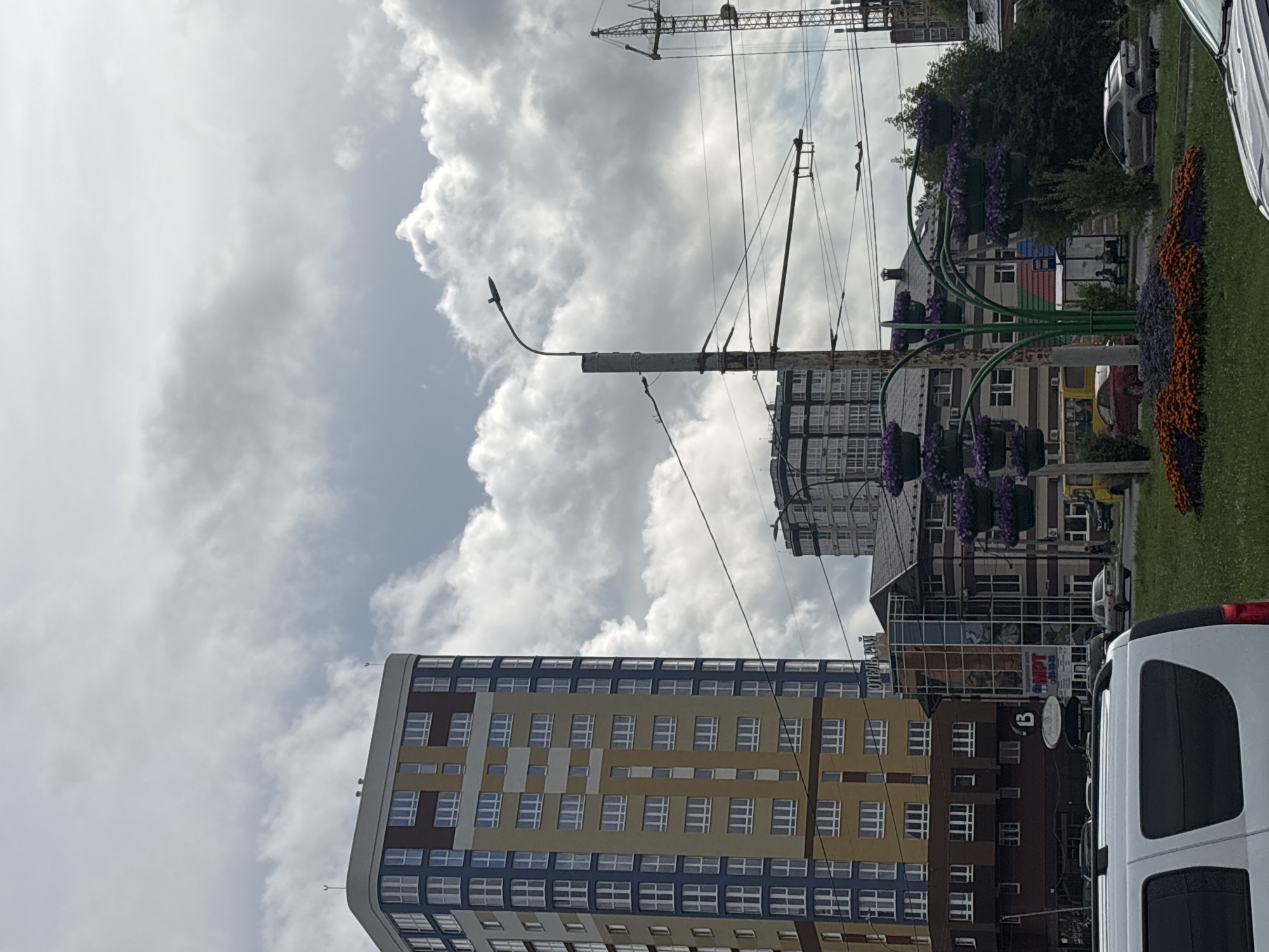 This is a photograph taken from a street level perspective, looking upwards at a cityscape with a predominantly cloudy sky. In the foreground, the top of a white van is visible, obscuring the lower portion of the scene. To the left, a tall, multi-story residential building stands out, characterized by its distinctive color blocking of blue, yellow, and brown sections. The building features numerous windows with white frames.
Further into the image, a street lamp on a metal pole is centrally positioned. Overhead wires crisscross the frame, suggesting an urban environment with public transportation or power lines. Behind the street lamp, another tall building with a dark facade and many windows is visible. To the right, a construction crane is partially visible against the sky, indicating ongoing development.
In the middle ground, a multi-story commercial building with a glass facade and a sloped roof is situated. Below it, a street-level view reveals storefronts, with one sign reading "MPT". In front of this building, a decorative structure resembling a tree is adorned with numerous planters filled with purple flowers. A bed of orange and purple flowers is also planted at the base of this structure.
Several cars are parked or driving on the street, including a silver sedan and a red sedan. A yellow bus is also visible. The environment appears to be a street in the city of Ivanovo, Russia, with elements of both residential and commercial architecture, along with infrastructure like power lines and a construction crane. The presence of the decorative flower display suggests an effort towards urban beautification.