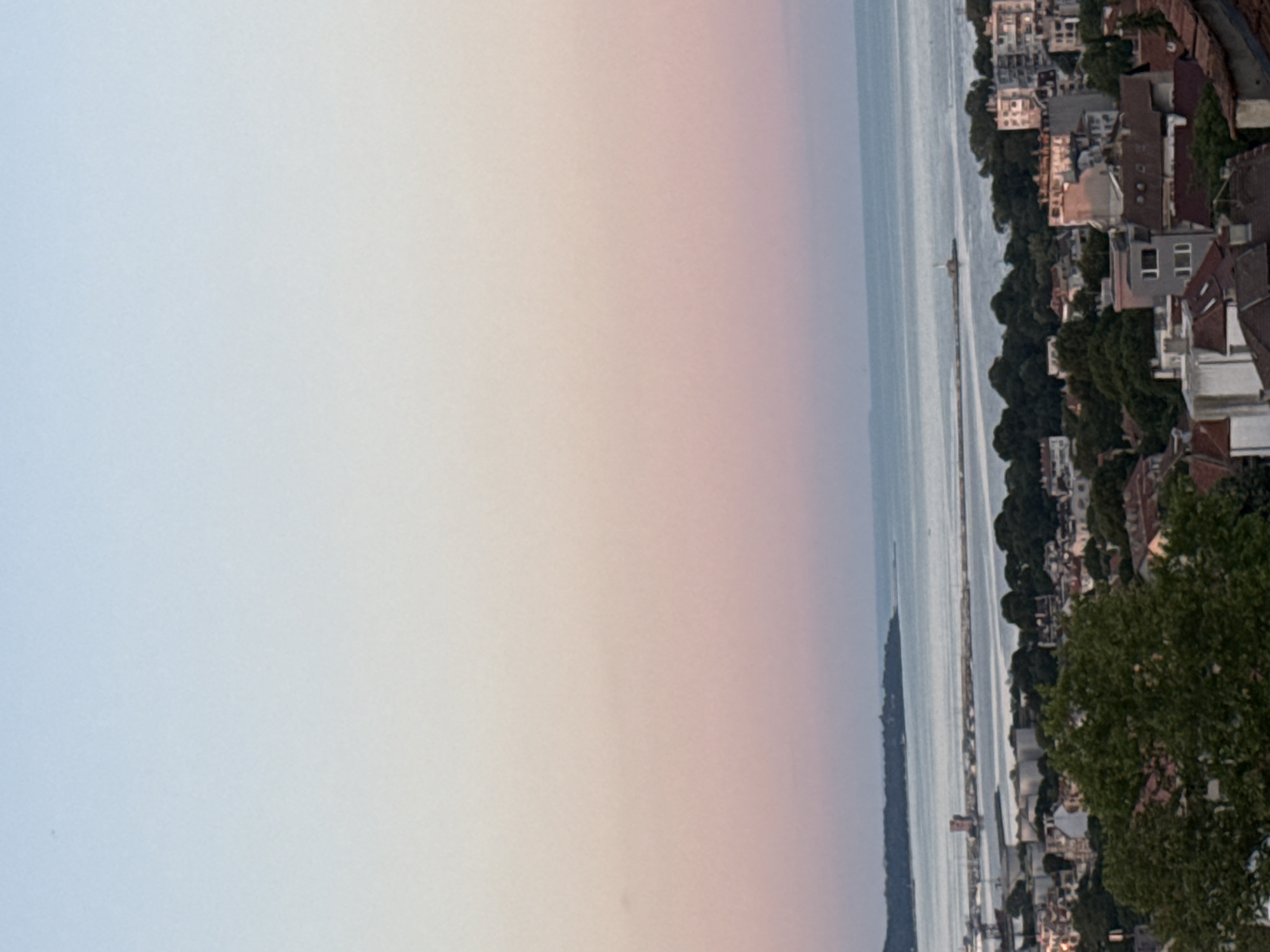 This is a wide-angle, high-angle, eye-level shot of a coastal city during either dawn or dusk. The sky transitions from a pale blue at the top to soft pinks and peaches closer to the horizon. Below the sky is a wide expanse of calm, pale blue water. In the distance, a thin, dark line of trees runs along the coast, separating the water from the city. A man-made structure, possibly a pier or breakwater, extends into the water from the left, with a tower-like structure visible on it. Further to the right, a small white lighthouse or beacon stands on a similar structure. The city itself is characterized by a dense collection of buildings with red-tiled roofs and some taller, modern structures. Lush green trees are interspersed throughout the urban landscape. The overall impression is one of a serene coastal settlement at the edge of the day.