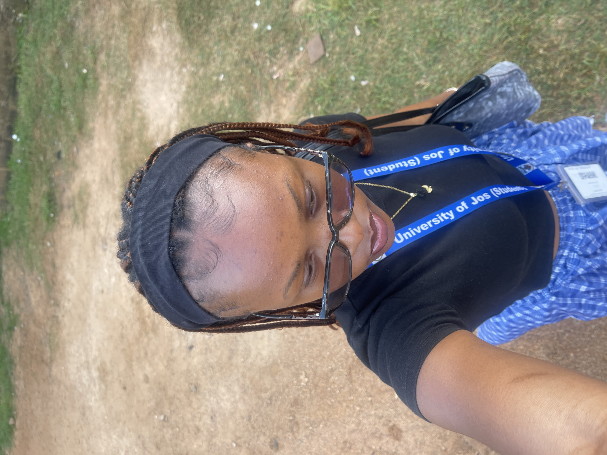 A young woman with braided hair, wearing a black headband, sunglasses, a black t-shirt, and blue plaid shorts, stands outdoors. She is wearing a blue lanyard that reads "University of Jos (Students)" around her neck, and carries a black and grey patterned bag. The ground is a mix of dirt and dry grass, with some green grass visible in the background. The lighting suggests it is daytime.
