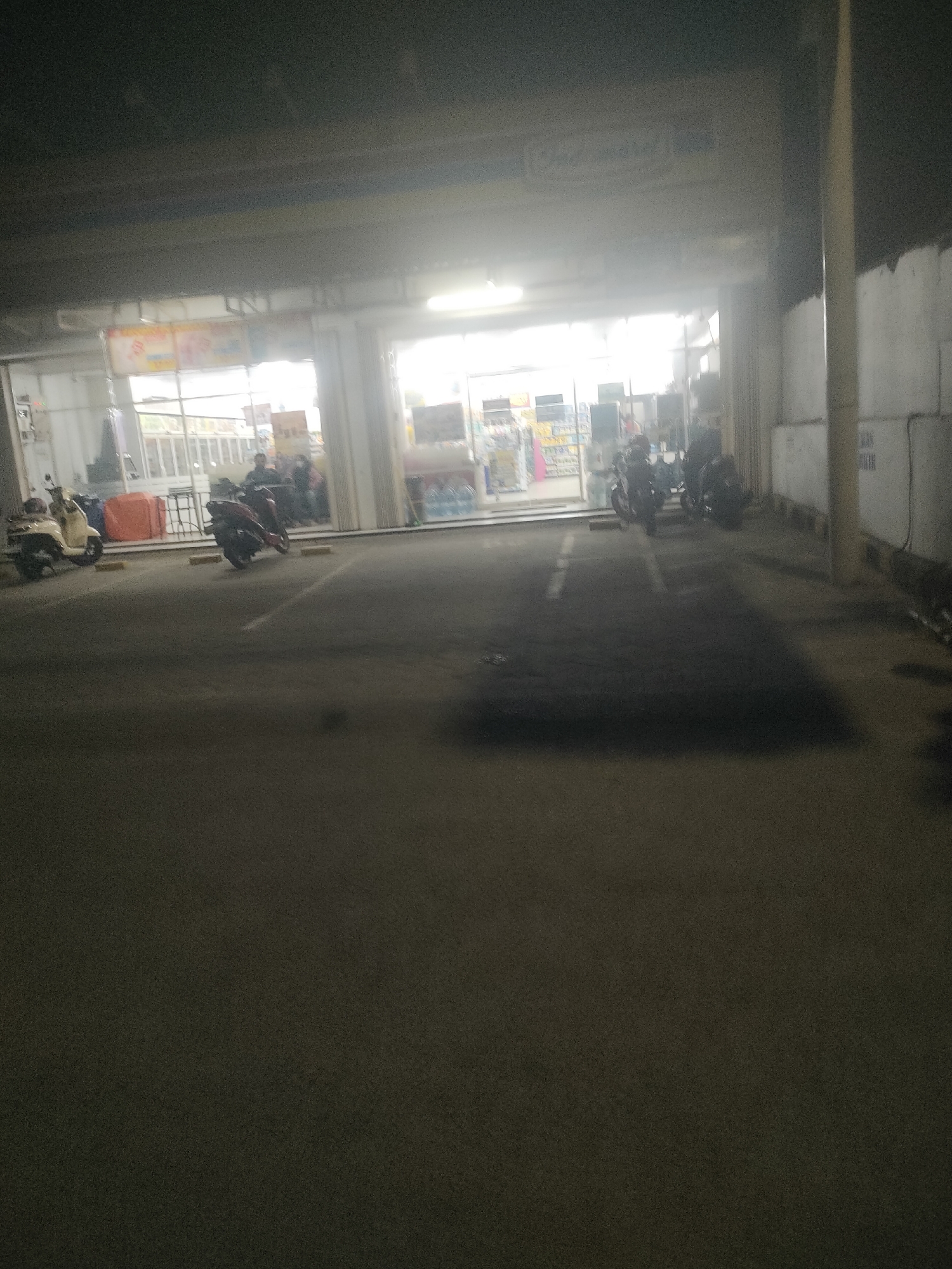 A dimly lit exterior view shows a paved area in front of a storefront. Several motorbikes are parked on the paved area. In the background, a glass-enclosed storefront is visible, with lights illuminating the interior. Figures of people can be seen inside the store and a few figures are standing in the doorway. The scene appears to be outdoors at night, possibly in a parking lot. The location is identified as Jogosatru, Indonesia.