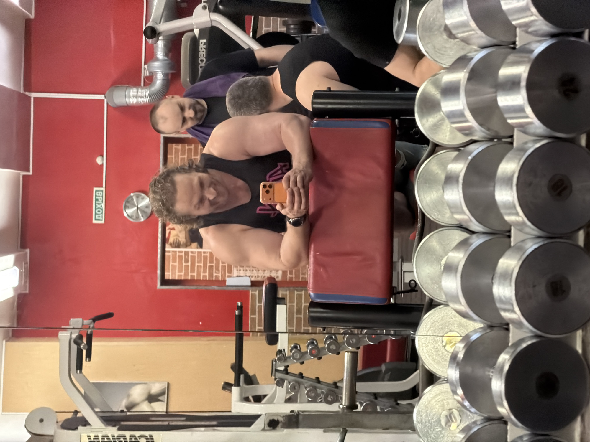 This image captures a scene inside a gym located in Saint Petersburg, Russia. In the foreground, a rack of dumbbells is visible, with their circular weights filling the bottom of the frame. Behind the dumbbells, a man with curly brown hair and a black tank top is leaning on a red and blue padded bench. He is holding an orange smartphone and appears to be taking a selfie or recording a video. His facial expression is one of amusement. To his right, another man with short dark hair and a purple towel draped around his neck is looking down. Further in the background, a third man in a black tank top is visible from behind, and parts of gym equipment are present. The gym walls are painted red, and a mirror reflects some of the equipment and people. A sign with Russian text, "ВХОД" (Entrance), is visible on the red wall. The overall impression is a candid moment captured in a functional gym environment.