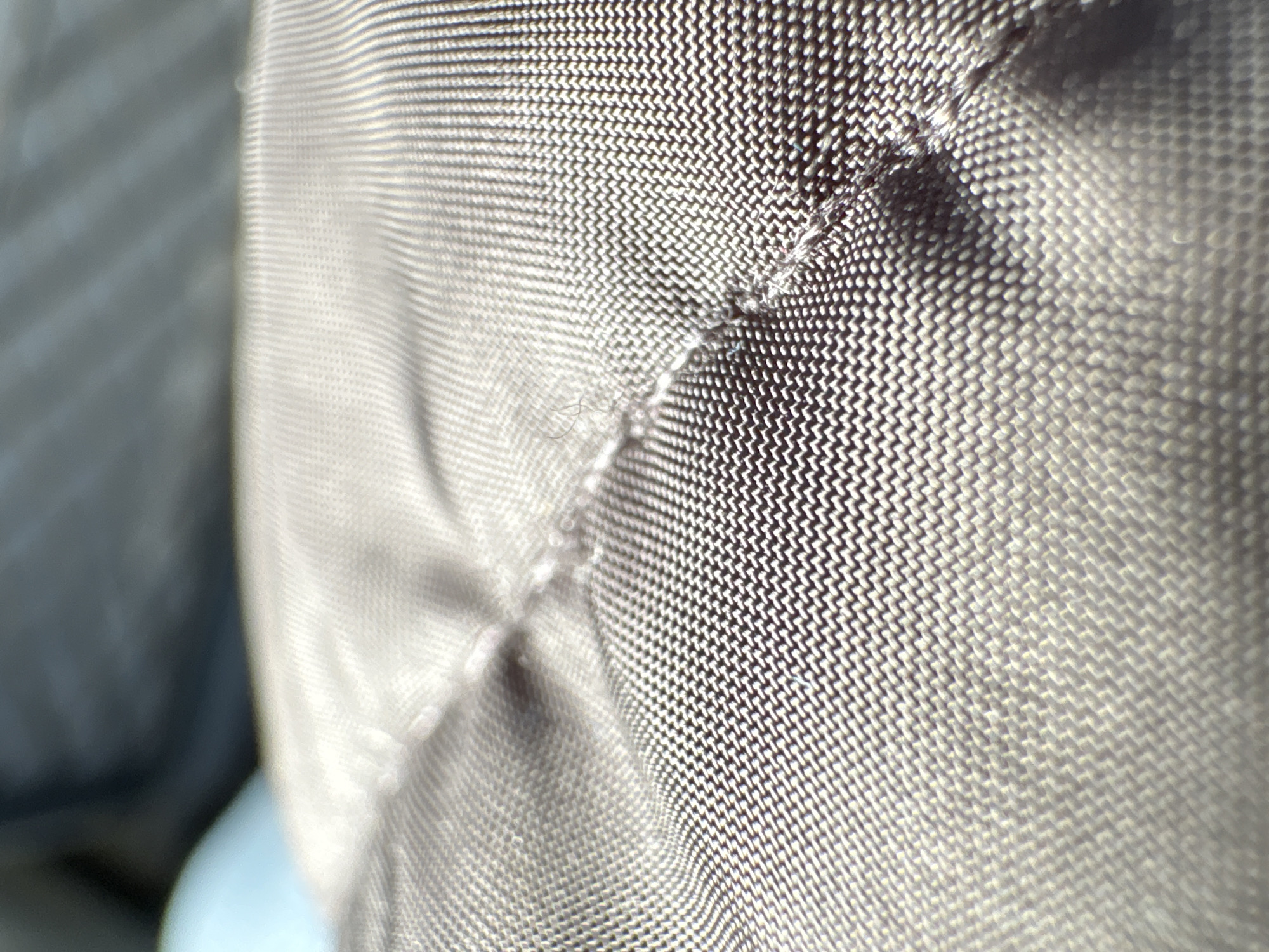 The image shows a close-up of a textured, woven fabric, possibly nylon or a similar synthetic material. The fabric appears to be a light gray or beige color, with a distinct weave pattern visible. The lighting is soft and even, casting subtle shadows that accentuate the texture of the fabric. The fabric is folded or draped, creating a small wrinkle or indentation near the center of the frame. The background is slightly blurred and out of focus. There is no indication of the location being Todoretsite, Bulgaria, or any people, actions or interactions.