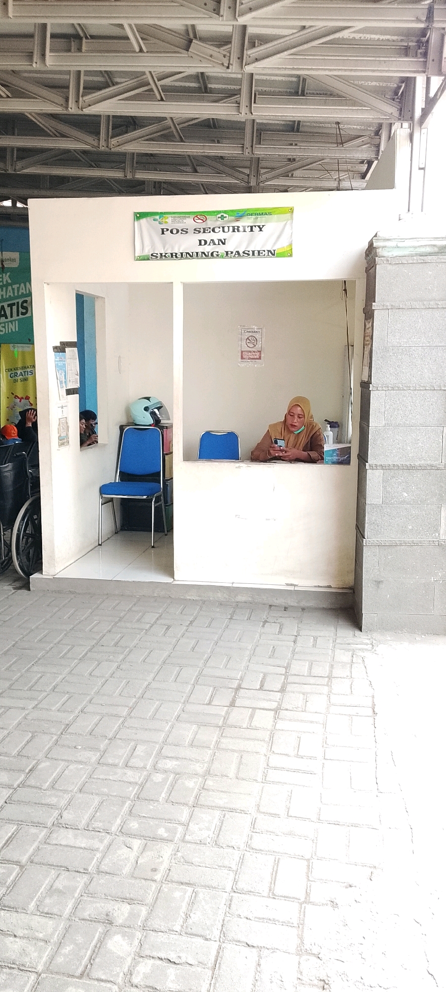 In Karangsatria, Indonesia, a person wearing a light brown hijab and a matching shirt is seated behind a counter inside a small security post. They are looking down at their phone, which they are holding in their hands. A blue chair is placed in front of the counter. To the left of the counter, another blue chair is positioned. A light blue helmet rests on a shelf behind the counter. A sign with Indonesian text and a red prohibition symbol is visible on the wall behind the person. The security post is located outdoors, with a paved walkway in front of it. The walkway is made of rectangular grey paving stones arranged in a patterned fashion. A section of a building with exposed metal beams is visible in the background. The overall scene suggests a functional area, possibly at a health clinic or similar facility, where security and patient screening are conducted.