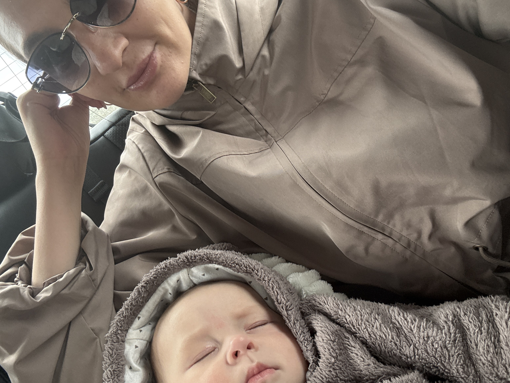 A woman wearing sunglasses and a light brown jacket is positioned above a sleeping baby wrapped in a grey blanket with a hooded section, likely inside a car. The location is presumed to be Sochi, Russia. The woman's hand is near her face, and the scene is well-lit.