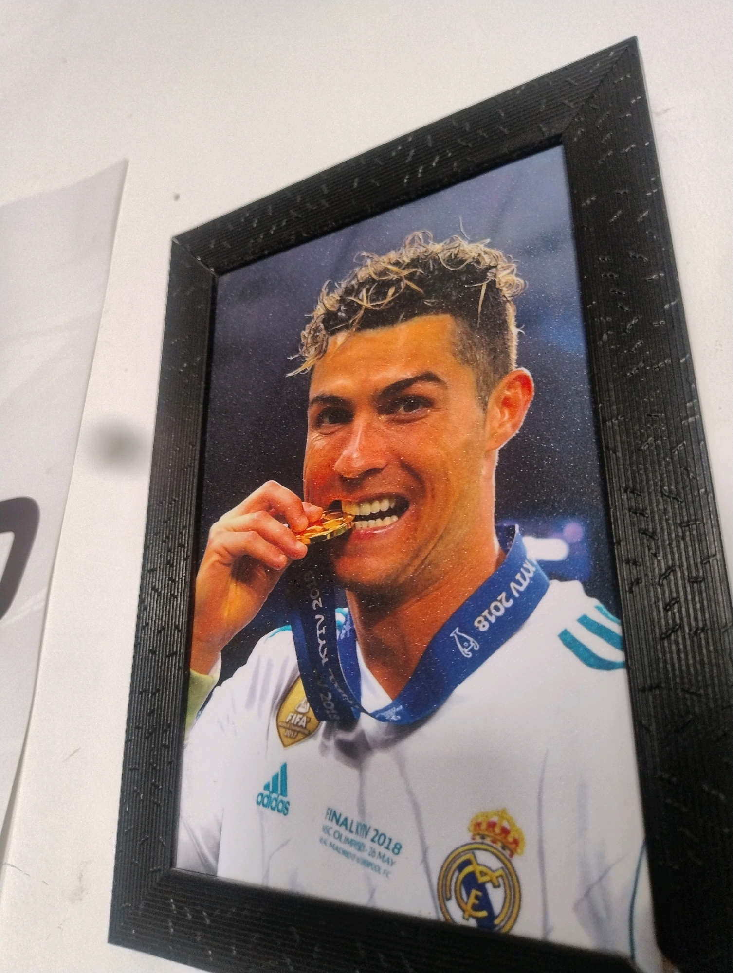 This is a photograph of a framed portrait of Cristiano Ronaldo, a professional footballer, biting on a gold medal. He is wearing a white jersey with blue stripes on the shoulders, a blue ribbon with text "KYIV 2018" and "FINAL 2018" printed on it, and the Real Madrid crest. He has curly hair and is smiling. The photograph is taken indoors, possibly on a wall, given the presence of another white, slightly angled surface to the left, which could be a poster or another wall. The overall lighting suggests an indoor setting. There is no visible information indicating the location in Bara, India.