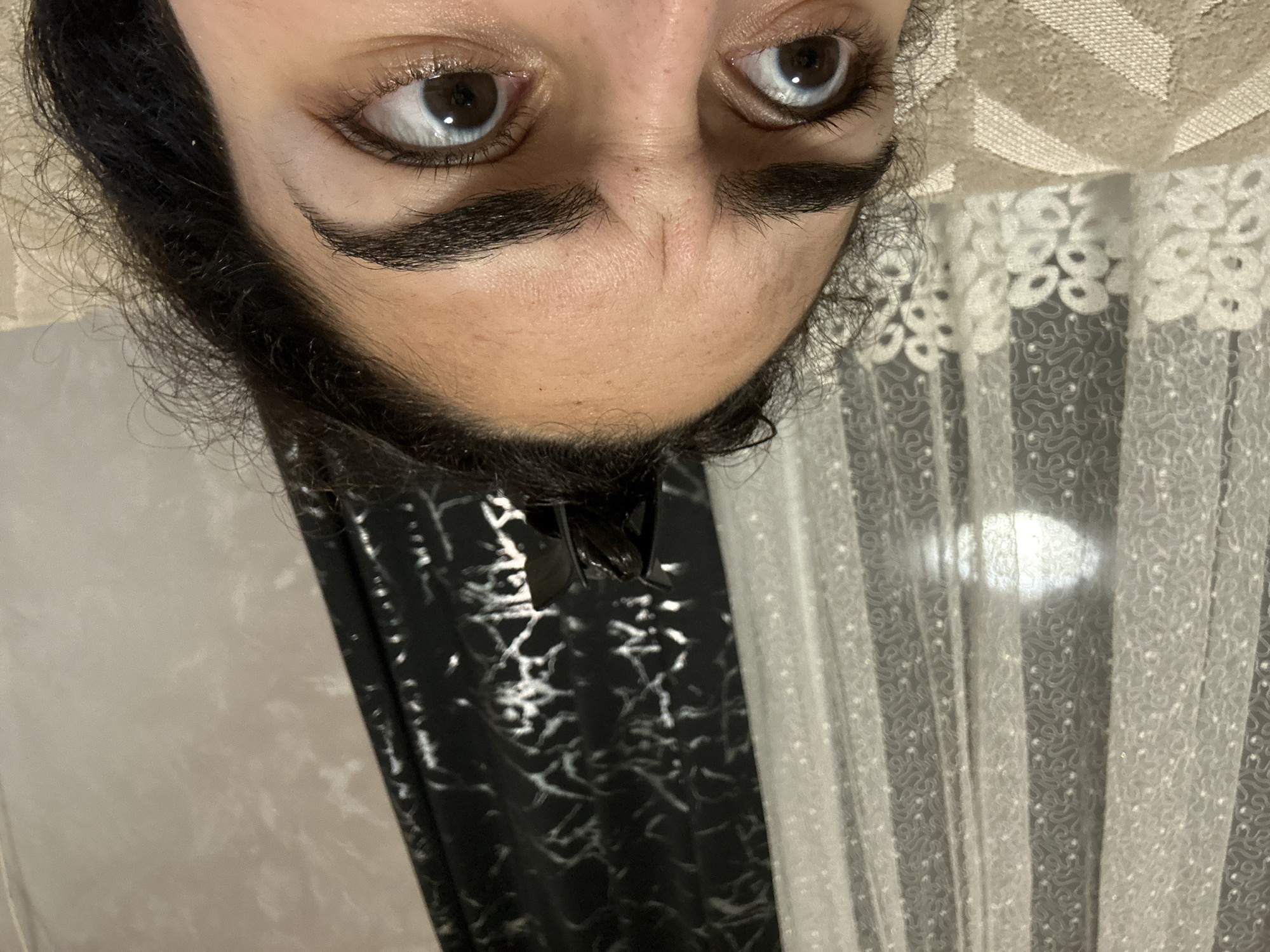 The image shows a person's face from a low, upside-down angle. The person has dark hair and dark eyebrows. The eyes are brown. The background includes a white curtain with a patterned design and a black fabric with silver patterns. The location is not discernible from the image.