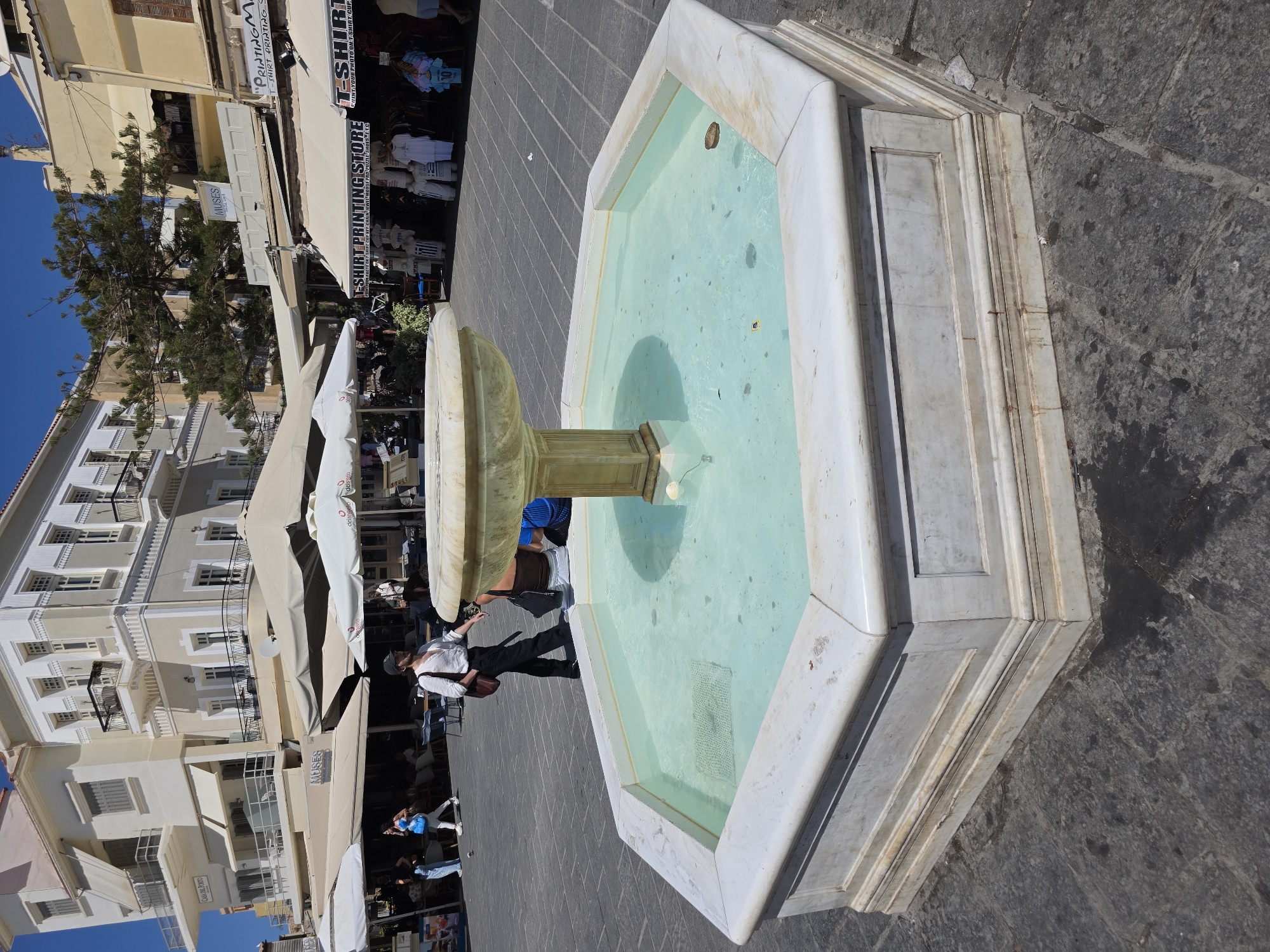 An outdoor, octagonal, marble fountain with light blue water is the central focus of the image. The fountain is situated on a grey, stone-tiled plaza. In the background, there are several buildings with light-colored facades and awnings, suggesting a commercial area. Shops with signs such as "Muses" and "T-SHIRT PRINTING STORE" are visible, indicating a tourist or shopping district. People are present in the scene, including a person wearing a white shirt and black pants walking towards the foreground, and others seated or standing in the background near the shops and under umbrellas. The sky is clear and blue, suggesting a sunny day. The overall environment appears to be a public square in a Mediterranean city, likely Chania, Greece, given the architectural style and presence of shops catering to tourists.