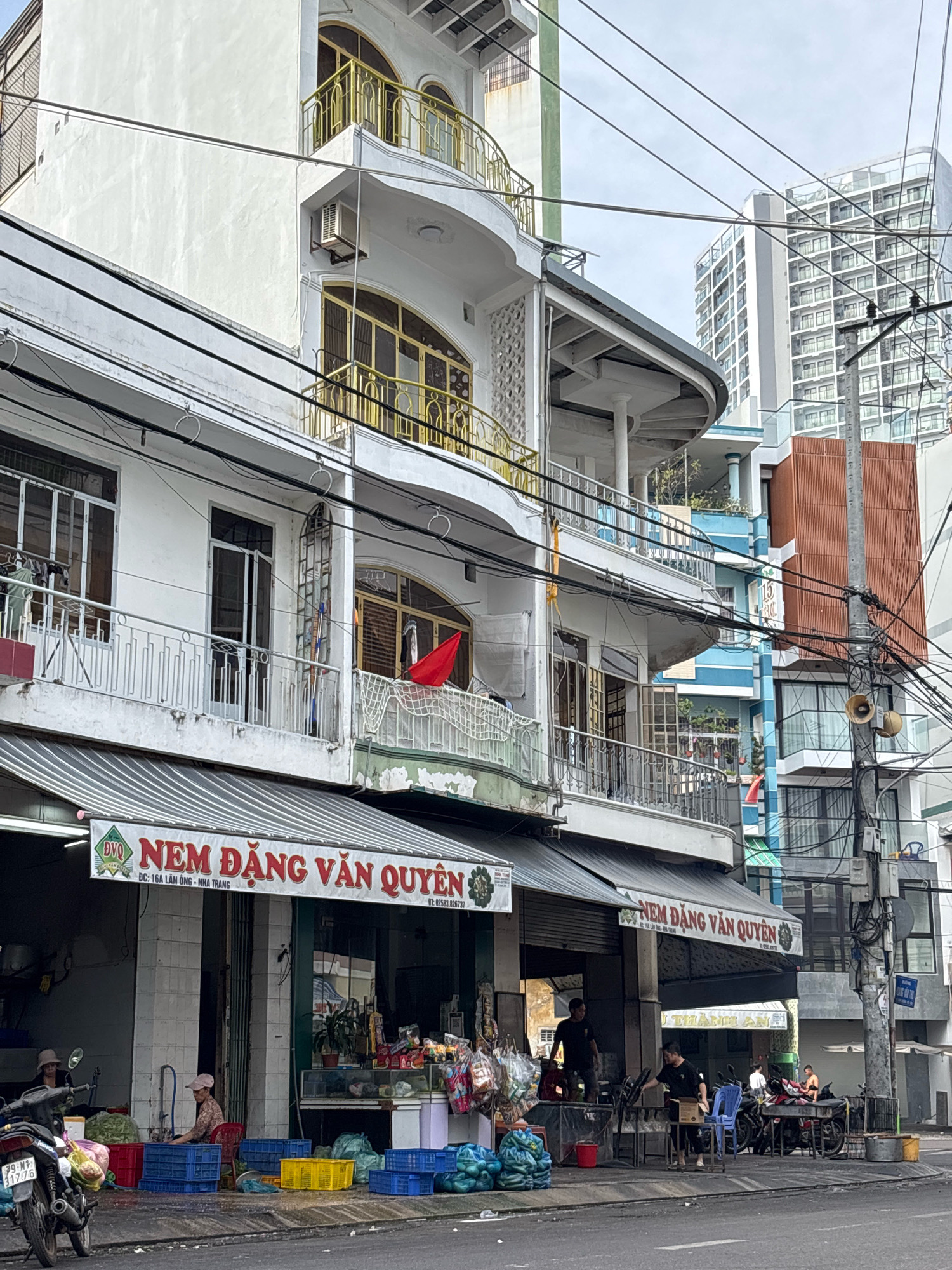 The image depicts a street scene in Nha Trang, Vietnam. In the foreground, a street-level shop with an awning displays a banner reading "NEM ĐẶNG VĂN QUYỀN" and an address "DC: 16A LẦN ÔNG - NHA TRANG". Several people are visible in and around the shop, some appearing to be customers and others working. The shop sells produce, with crates of vegetables and bags of goods displayed on the sidewalk. Motorbikes are parked along the street. Above the shop, a multi-story building features balconies and windows with ornate yellow railings. The background shows taller modern buildings, including a high-rise apartment complex, and a tangled network of electrical wires. The sky is overcast.