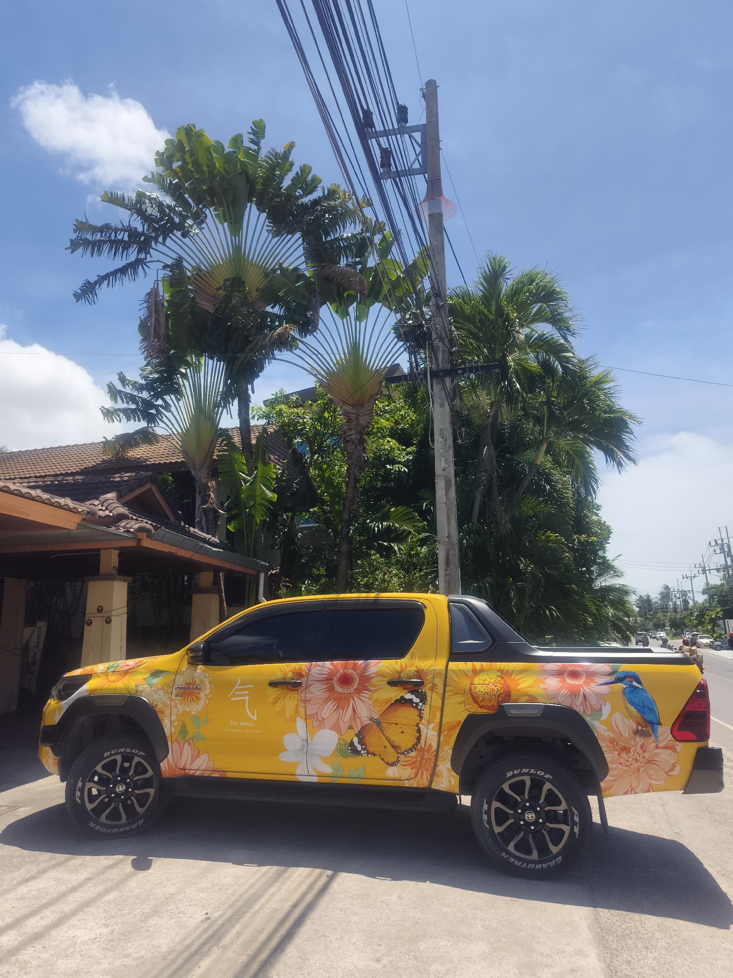 A bright yellow pickup truck, vibrantly decorated with floral and fauna designs, is parked on a concrete surface alongside a road in Baan Bang Rak, Thailand. The truck's design includes sunflowers, butterflies, and a blue bird. The surrounding environment features a building with a tiled roof and support columns, tall trees that appear to be traveler's palms, other lush tropical foliage, and a utility pole with multiple wires. The sky is blue with scattered clouds. Cars can be seen in the distance on the road.