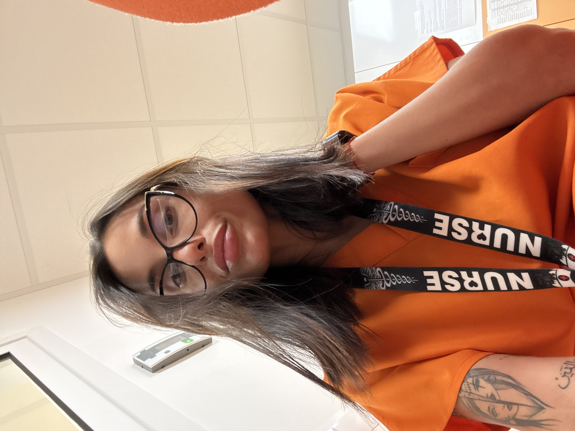 A young woman with dark hair and black-rimmed glasses is taking a selfie indoors, likely in a hospital or clinic setting. She is wearing an orange scrub top and has two lanyards with the word "NURSE" printed on them hanging around her neck. A tattoo of two faces is visible on her left arm. The background includes a white ceiling with fluorescent lights, a white wall with a light fixture, and a window. There are also papers visible on a surface to the right. The image does not provide any specific details about the city of Pleven, Bulgaria.
