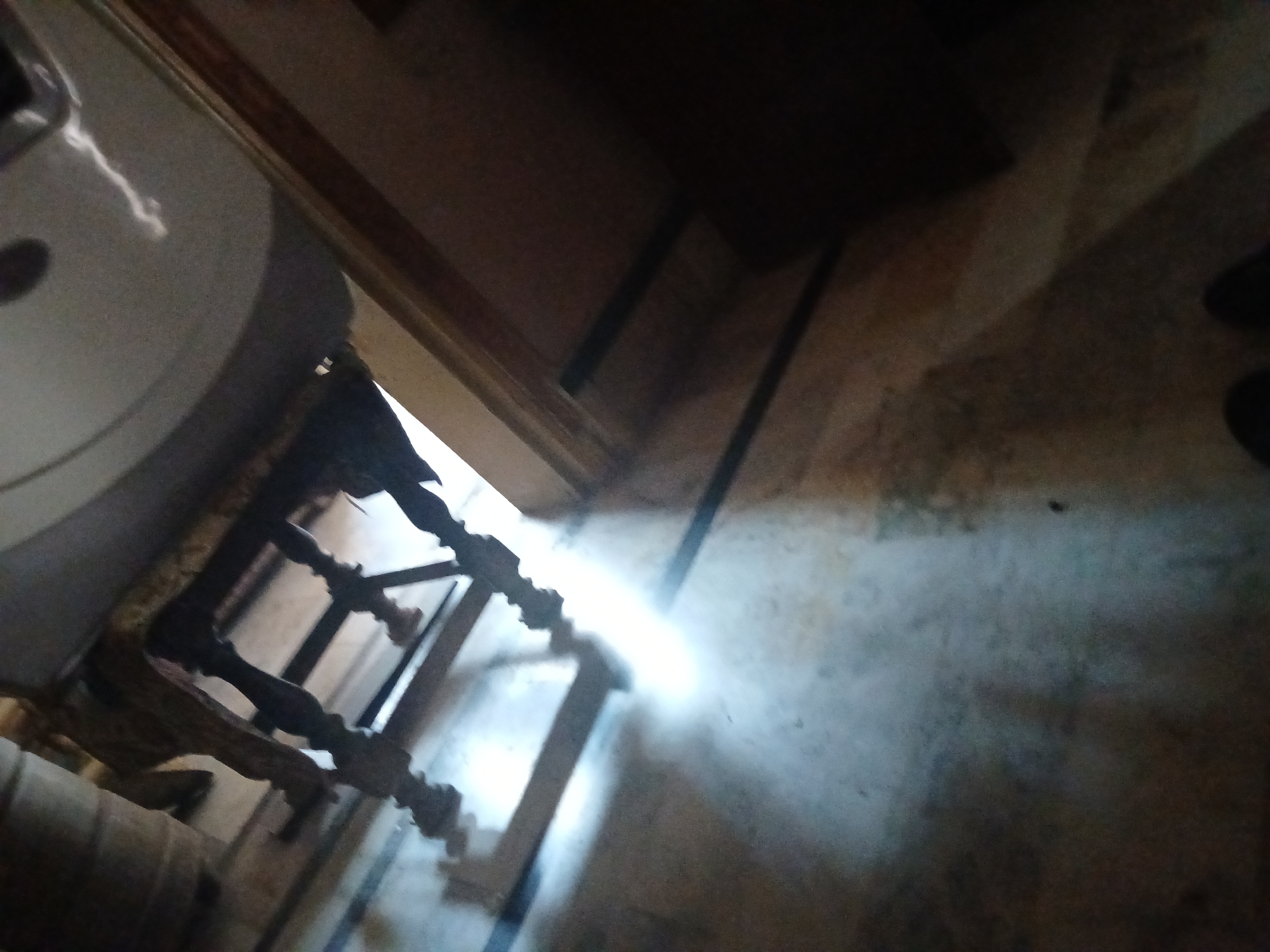 The image shows an unusual perspective, looking upwards at the underside of a wooden structure and a circular object above it. The wooden structure appears to be part of furniture, possibly a table or a bench, with decorative turned legs. Light streams from the left, illuminating a section of a concrete floor with a rough texture, marked by dark linear shadows cast by the wooden structure. The environment suggests an indoor setting, though the exact location and purpose of the structure are unclear. The image composition is abstract due to the extreme upward angle and the play of light and shadow. The overall impression is one of mystery and disorientation, with the bright light creating a focal point amidst the darker, more ambiguous areas. No specific location in Peshawar City Tehsil, Pakistan can be identified from the image.