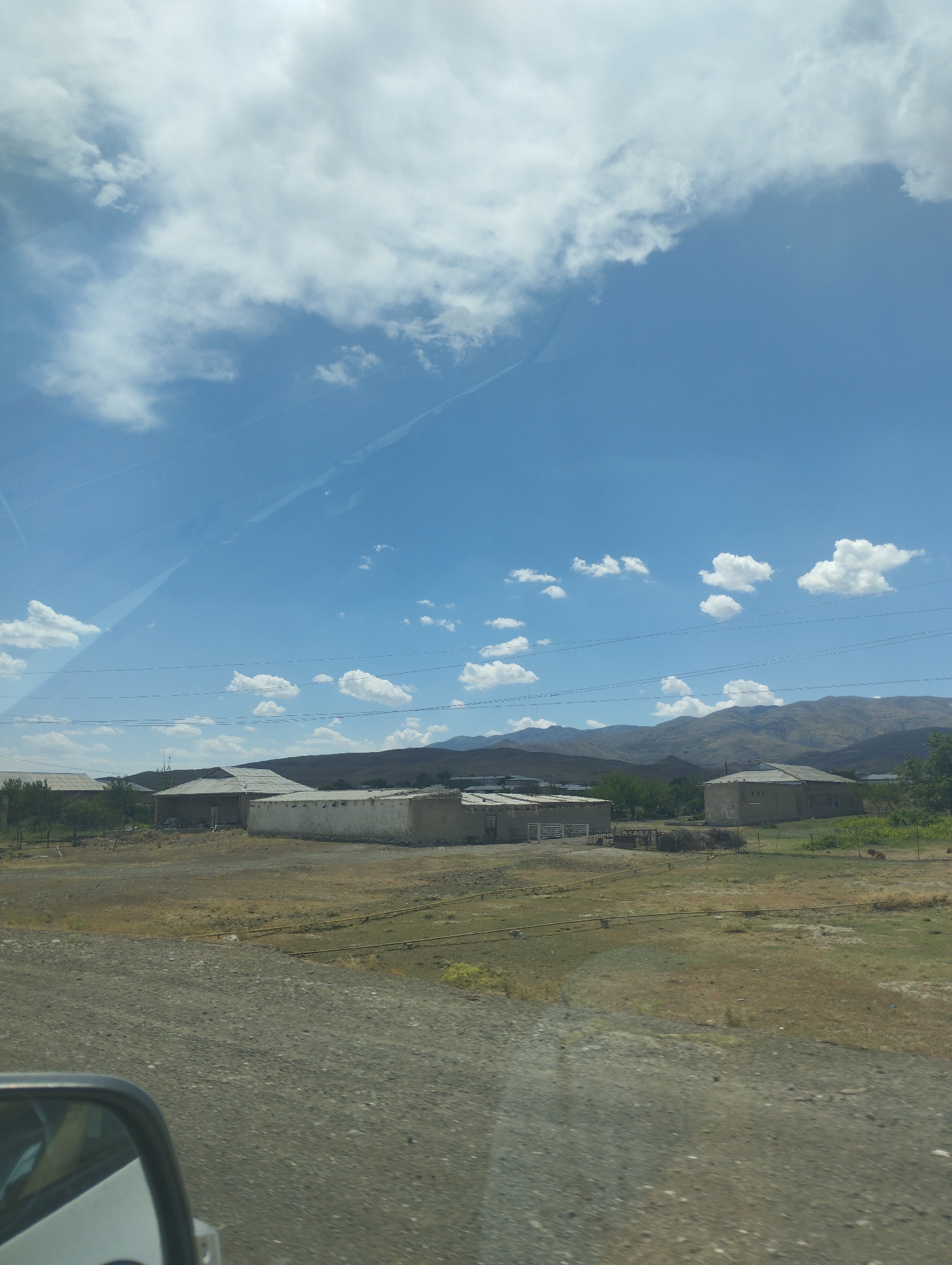 The image depicts a rural landscape in Asmansay, Uzbekistan, under a partly cloudy sky. In the foreground, a dirt road is visible, with the side mirror of a vehicle in the lower left corner. Beyond the road, there are several low-lying buildings, some appearing to be agricultural or storage structures with light-colored walls and corrugated roofs. The ground is covered with dry grass and scattered patches of dirt. In the middle ground, there are more of these simple structures and some trees. The background features rolling hills and mountains under a wide expanse of blue sky dotted with white clouds. There are no people visible in the frame, and the scene appears calm and undisturbed. The overall impression is one of a somewhat arid, rural environment.