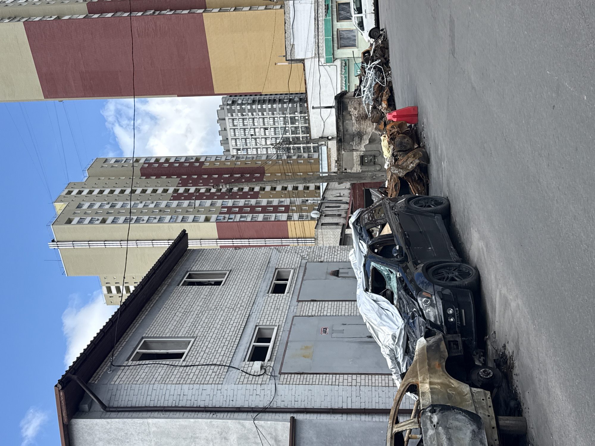 In an urban environment in Kyiv, Ukraine, two damaged vehicles are parked on an asphalt surface. The vehicle on the left is severely burned, with its rear end heavily charred and its wheels missing. It is propped up by wooden posts. The vehicle on the right is a black SUV, partially covered with a reflective tarp. Its windshield is shattered and its front end appears damaged.
Behind the vehicles, a low-rise brick building with several garage doors and windows is visible. Further in the background, several tall residential buildings dominate the skyline under a cloudy sky. To the right of the brick building, a pile of debris, including burnt metal and other scrap materials, is visible. A white van is parked in front of another low-rise building to the far right. The scene suggests a recent event of destruction or damage in the area.