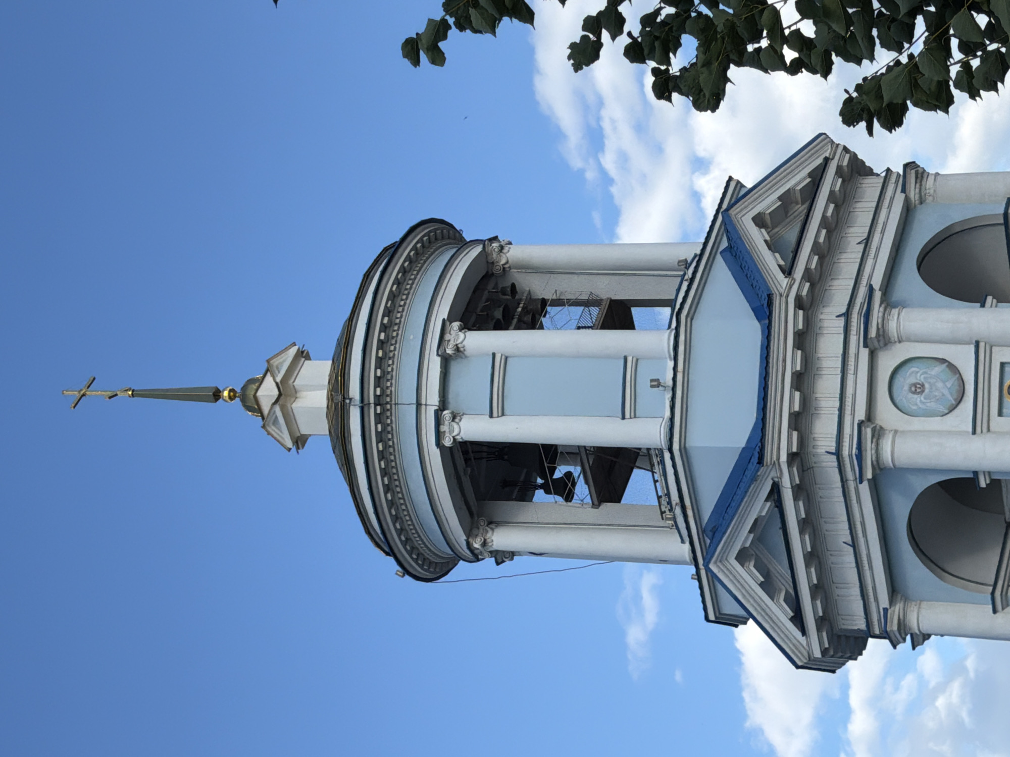 An upward, low-angle shot features the top portion of a light blue and white bell tower against a clear blue sky with scattered white clouds. The tower has a tiered, octagonal design. At the very top is a slender, gold cross atop a pointed spire, which sits on a golden dome. Below this is a circular structure with white columns, housing several dark bells visible through the open sides. Further down, a section of the tower has blue, angled roofing. At the base of the visible structure, there are arches and decorative moldings. To the right of the frame, dark green foliage from a tree encroaches on the scene. The image appears to be of the bell tower of the Church of the Transfiguration in Novaya Usman', Russia.