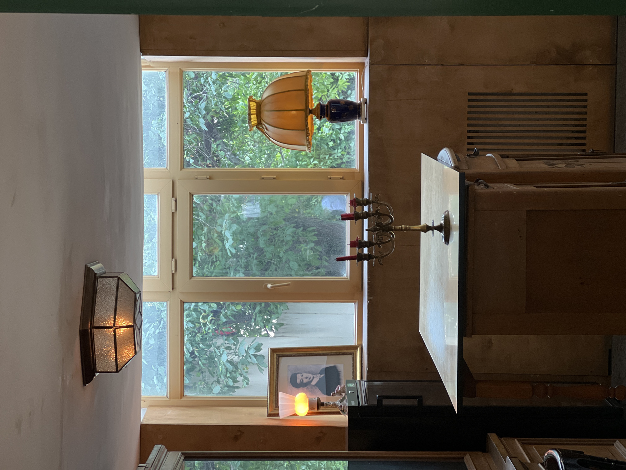 The image displays a warm, cluttered interior room in Yerevan, Armenia, captured through a vertically oriented photograph. A prominent light-colored, multi-paned window occupies the central space, looking out onto a dense expanse of vibrant green foliage, suggesting a lush garden or natural backdrop. The outdoor lighting indicates it is daytime, possibly overcast or late afternoon, as several artificial lights are on indoors.
On the wall to the left of the window, an illuminated wall sconce with textured, segmented glass and a dark metal frame emits a soft, golden glow. Above the window, perched on a narrow ledge, is a vintage-style table lamp featuring a fluted, amber-colored glass shade and a dark blue, ornate base, also illuminated.
The window sill below holds several decorative items. A brass-toned candelabra with three red, unlit candles stands near the right. To its left, a framed black and white portrait, depicting what appears to be a man with dark hair, is displayed. Beside the portrait, a small, fan-shaped decorative light casts an orange glow.
To the right of the window, the wall is covered with wooden paneling, which includes a dark, horizontal vent. Below this, a section of a large wooden furniture piece, possibly a dresser or cabinet with visible ornate carvings, is present. Its reflective top surface partially mirrors the surroundings. The lower foreground includes dark furniture and various other objects, such as a picture frame and a metallic item.
The overall impression is one of a lived-in, cozy space with a blend of natural light and warm, artificial illumination, creating a somewhat nostalgic and inviting atmosphere. No visible text is present in the image.