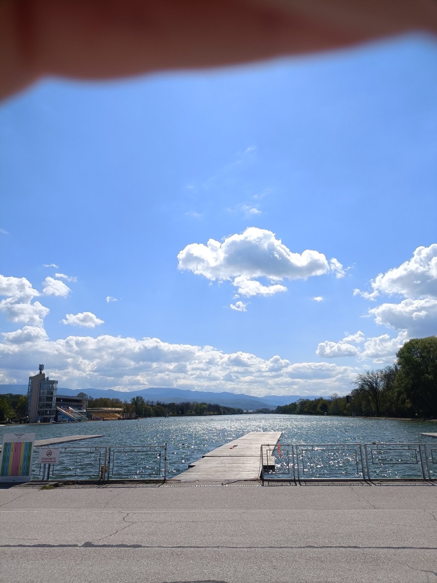 This image presents a broad view of a regatta venue under a bright, sunny sky. The primary subject is a wide body of water, likely a rowing canal or lake, which stretches into the distance, reflecting sunlight in shimmering patterns. A long, wooden pier extends into the water from the right side of the frame.
In the foreground, a paved area with metal railings separates the viewer from the water. To the left, a large information sign displays "Трасета на гребния канал" (Tracks of the rowing canal) at the top, followed by colored stripes indicating lanes. Below this, a no-jumping symbol explicitly states "СКОКОВЕТЕ ЗАБРАНЕНИ" (Jumping forbidden).
On the left bank of the canal, a modern, multi-story building with a distinctive tower-like section is visible, suggesting a control tower or part of a grandstand complex. Beyond this, along both banks of the canal, lush green trees line the water's edge. In the far distance, a range of mountains rises under a clear blue sky adorned with scattered white cumulus clouds.
The scene depicts a peaceful, serene environment, indicative of daytime with bright sunshine. No specific activity or people are present, but the infrastructure strongly suggests a venue for water sports like rowing or kayaking. The visible Cyrillic text and the architectural style are consistent with the location context of Plovdiv, Bulgaria, likely depicting the Regatta Venue. A blurry, reddish-brown obstruction, appearing to be a finger, is visible in the top-left corner of the image.