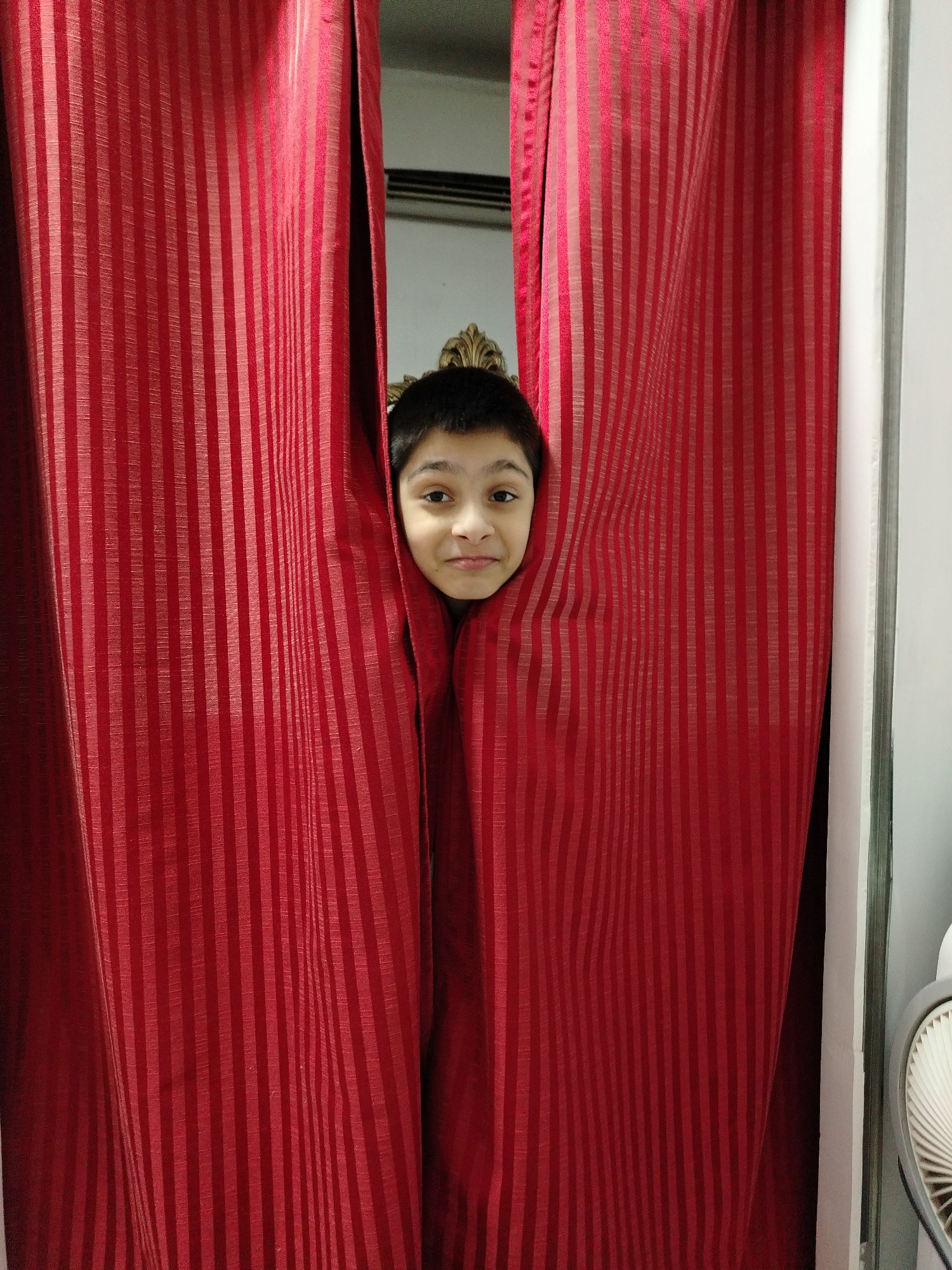 A child's face peeks through a gap in red striped curtains. The child has dark hair and is looking at the camera with a slight smile. The curtains appear to be hanging in a room with a white wall and a glimpse of what might be an air conditioning unit in the background. The image was taken in Al Mas'oudi, United Arab Emirates.