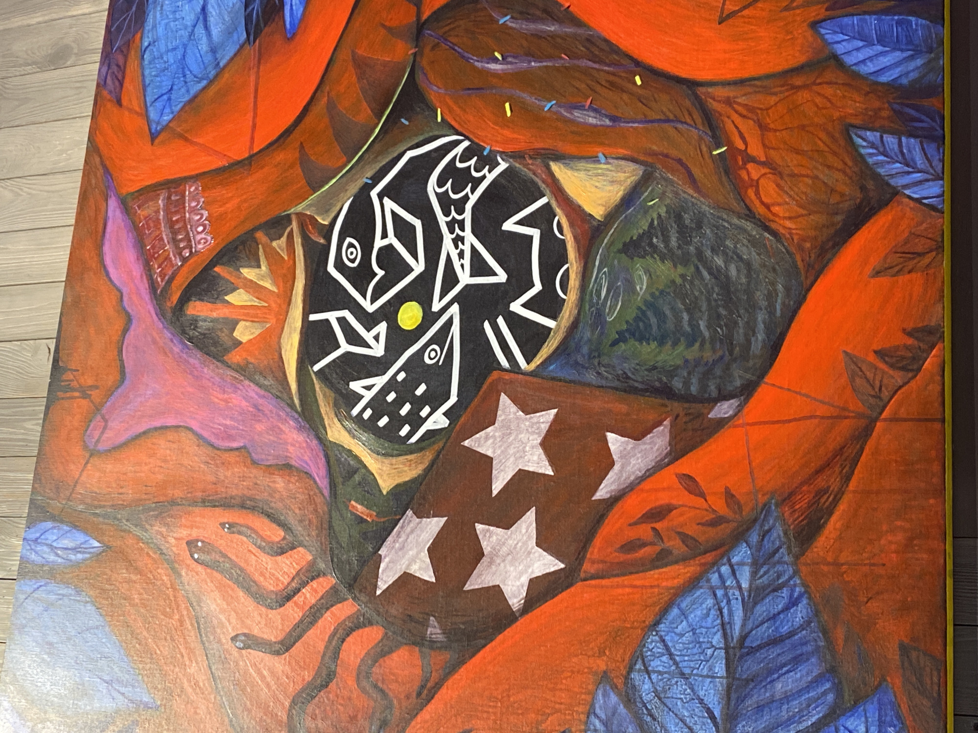 The image shows a painting with a chaotic arrangement of subjects and objects. The background is predominantly orange with blue leaves interspersed throughout. There is a black circular shape with white lines depicting fish and geometric patterns. Below it is a rectangular shape with three white stars on a brown background. To the left, there is a pinkish-purple shape. There are also brown, tentacle-like lines and dots. The painting is framed in yellow. The location of the painting and the city of Perm, Russia are not identifiable from this picture.