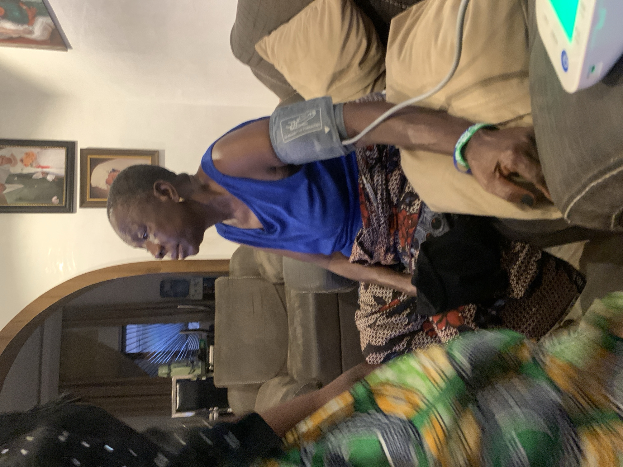 An elderly woman with short gray hair is seated on a brown sofa with tan cushions, a blood pressure cuff attached to her left arm. She wears a blue tank top and a patterned skirt or dress. The blood pressure machine is visible in the upper right. Framed portraits hang on the wall in the background. The environment appears to be a domestic setting, possibly a living room, with an arched doorway leading to another room. A blurry person wearing a green and yellow patterned garment is partially visible in the foreground. The location in the city of Jos, Nigeria, cannot be determined from the visual data.