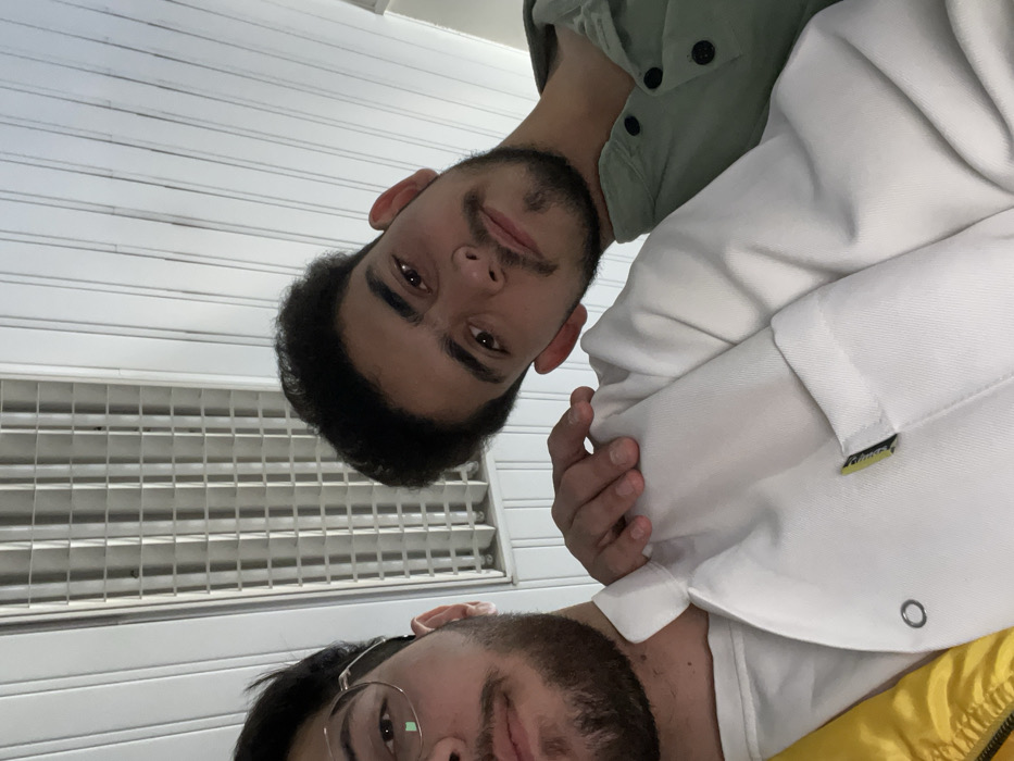 This image captures two young adult males in a close-up, low-angle shot, likely a selfie, taken indoors in Homs, Syria. The male on the left is partially visible, showing his left profile from the nose down, a dark beard, and round, light-framed eyeglasses. He wears a white t-shirt under a yellow garment, and a white collared jacket or shirt with a small yellow and black tag on a breast pocket, secured by a silver eyelet/rivet. The male on the right is centrally positioned, facing the camera with a direct gaze, displaying dark hair, a mustache, and a short beard. He wears a light green collared shirt with a placket featuring three dark buttons. His left hand rests on the left male's right shoulder. The overhead environment consists of a white or light-colored ceiling with parallel decorative strips and a rectangular fluorescent light fixture containing multiple tubes and a grid diffuser.