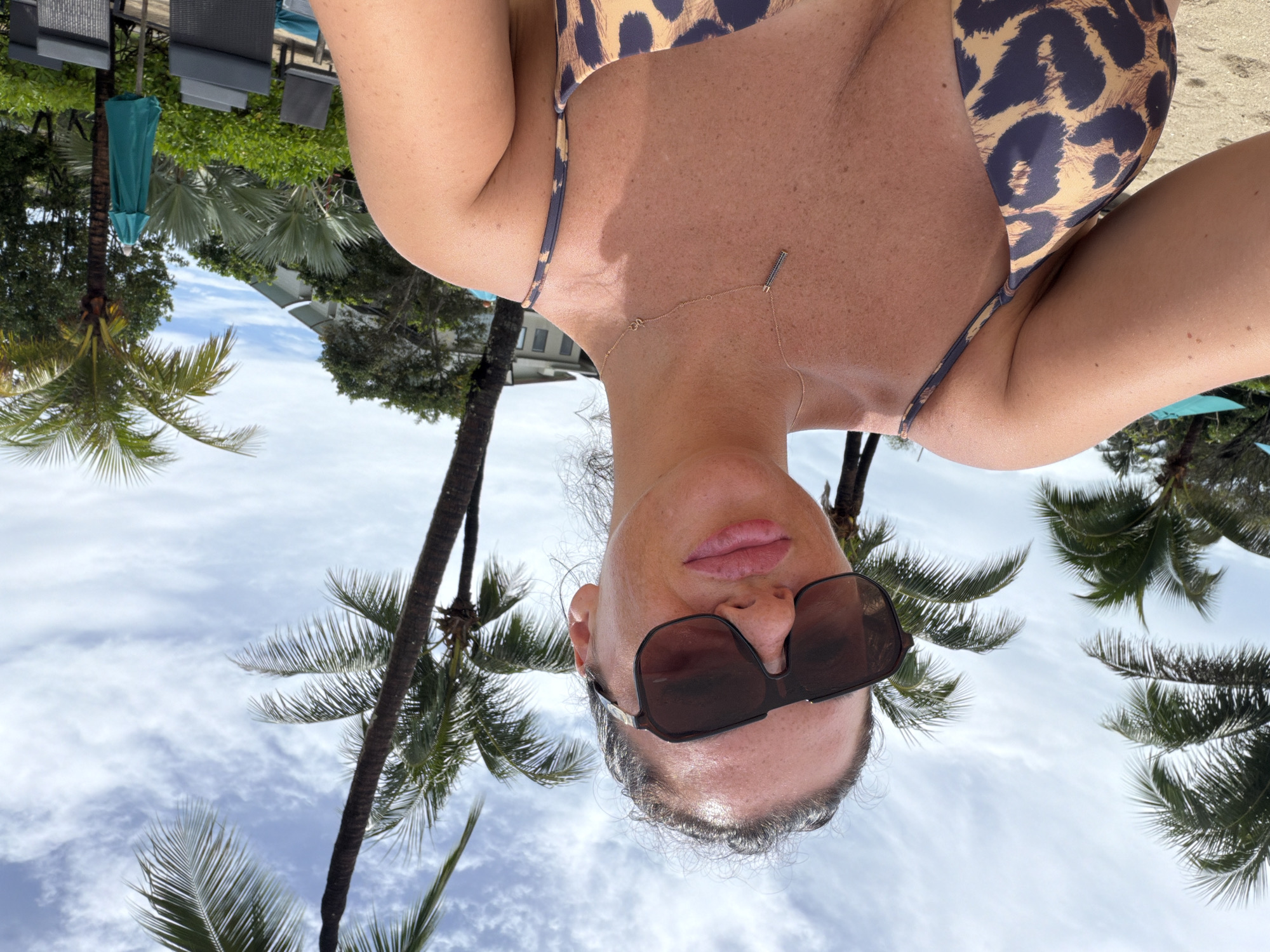 A woman is taking a selfie on a sunny day. She is wearing large sunglasses, a leopard-print bikini top, and a delicate necklace. The background features palm trees, a sandy beach, lounge chairs, and umbrellas. The overall setting suggests a tropical resort location, possibly in the city of Bang Muang, Thailand, given the presence of lush vegetation and a cloudy, bright sky.