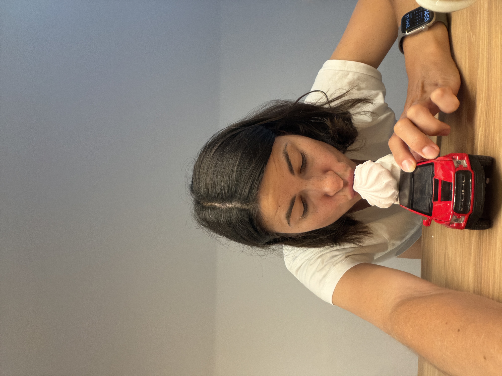 A person is depicted indoors, seemingly posing for a selfie. She is dark-haired and light-skinned, wearing a white t-shirt. She has a smartwatch on her wrist. She is positioned next to a red toy truck, and a swirl of white cream is placed between her face and the truck. The background appears to be a light gray wall and a light-colored wooden surface. There is no information available in the image to infer any location in the city of Budva, Montenegro.