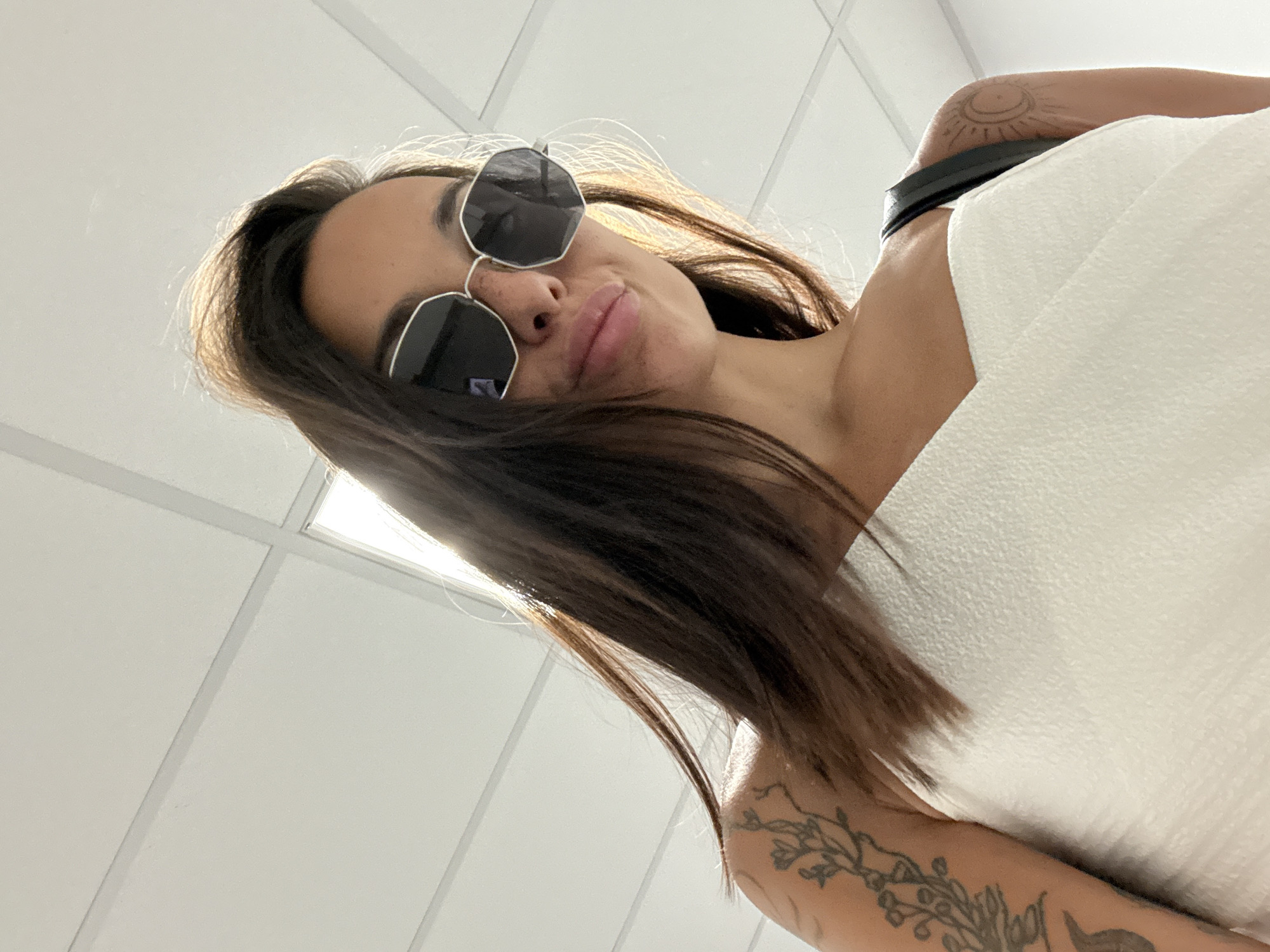 In Pleven, Bulgaria, a woman is pictured from a low angle, looking up towards a white ceiling with fluorescent lighting panels. She is wearing a white top and large, polygonal, dark-lensed sunglasses. Her long, dark hair is falling around her face and shoulders. She has tattoos on her left arm and shoulder, one appearing to be a floral design and the other a celestial symbol. The background is the interior of a building, likely illuminated by the overhead lights. The overall impression is a candid selfie taken indoors.