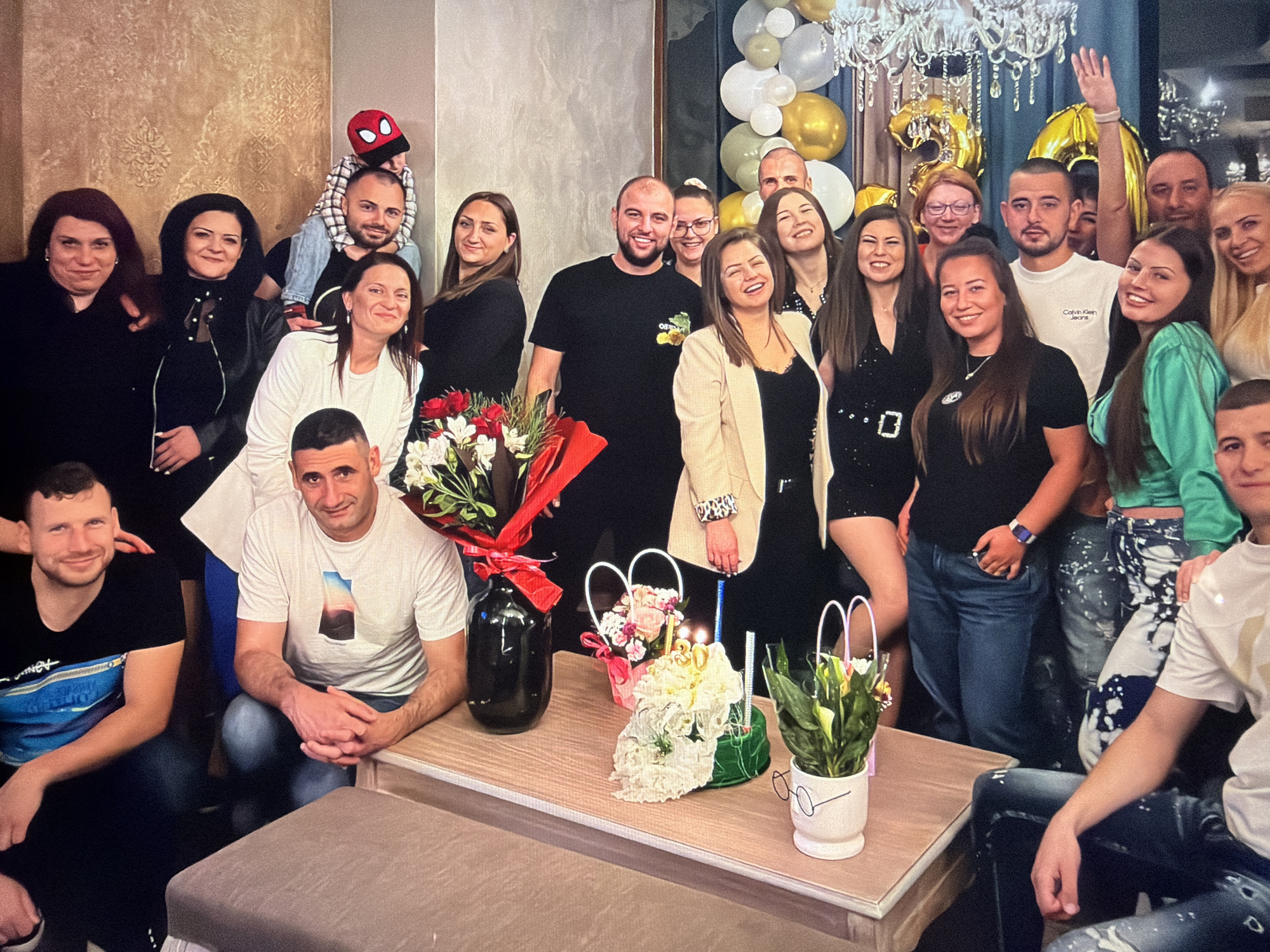 A group of approximately 20 people of varying ages are gathered indoors, seemingly for a celebration, possibly a 30th birthday, as indicated by the cake with the number "30" on it. The group is a mix of genders, with some individuals standing and others seated or partially visible at the edges of the frame.
At the center of the scene, a light-colored coffee table holds a cake decorated with white flowers, number candles, and several decorative floral arrangements. One flower arrangement is in a black jar tied with a red ribbon, another is in a white basket with a pink ribbon, and a third is in a white pot with glasses drawn on it.
The background includes light-colored textured walls, a glass-paneled section, and a chandelier. Gold and white balloons are visible near the glass panel, further suggesting a celebratory occasion. One person near the upper right of the photo is waving their hand.
The location is likely an indoor venue in Burgas, Bulgaria, given the request in the prompt.