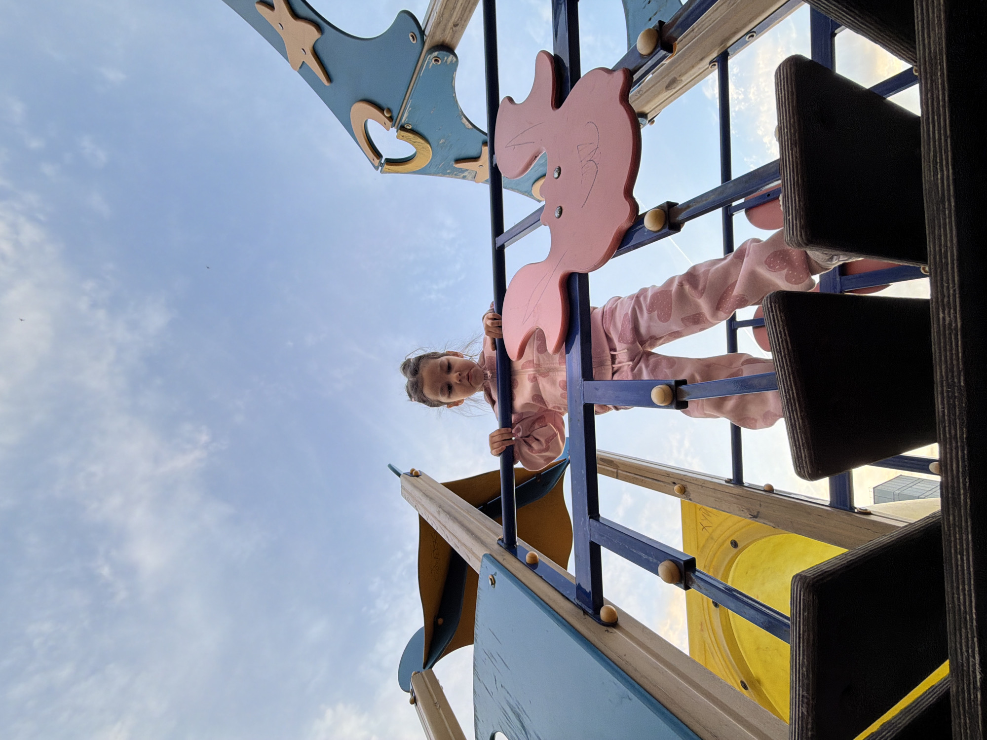 A low-angle shot depicts a young girl climbing on playground equipment in Chaikovsky, Russia. The girl is wearing a pink outfit and has her hair pulled back. The playground equipment consists of blue and yellow structures with wooden accents. In the background, a partly cloudy sky is visible.