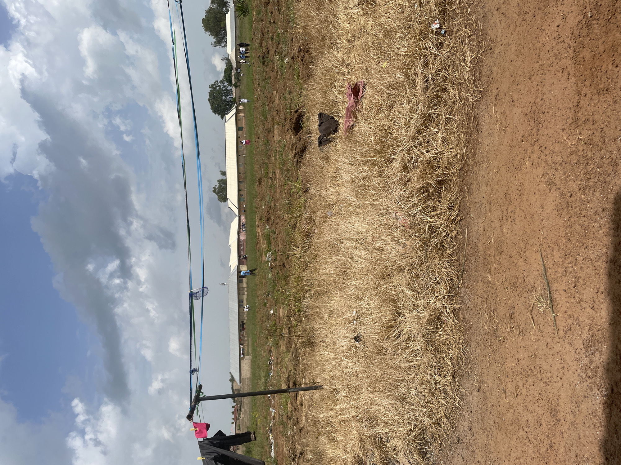 This media file captures an outdoor scene in Durumi, Nigeria, under a partly cloudy sky, suggesting it is daytime. The foreground features a clothesline with several items of clothing, including a bright pink garment and dark-colored pants, hanging from it. Beyond the clothesline, the ground transitions from dry, tall grass to bare, reddish-brown soil. In the mid-ground, there is a cluster of buildings, one of which appears to be a large, white tent structure, possibly a temporary shelter or event hall. Scattered around the buildings and on the ground are various pieces of debris.
Several individuals are visible in the mid-ground, some standing near the buildings and others walking. Their presence suggests some activity or gathering is occurring. A few trees are also present in the background, adding to the rural or semi-rural feel of the location. The overall impression is of a functional, perhaps somewhat makeshift, settlement or event site. The weather appears fair, with a mix of sun and clouds. No discernible text is visible in the image.
