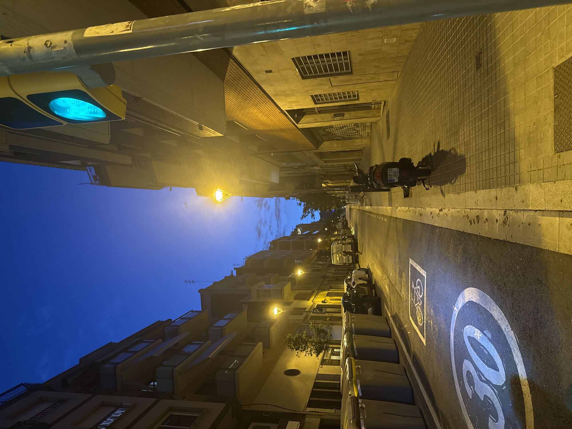 The image shows a street scene in Barcelona, Spain, at dusk or dawn, with the sky a deep blue. The street is lined with multi-story buildings featuring balconies and windows. A traffic light with a green signal is visible on the right side of the frame, attached to a metal pole. The sidewalk is paved with square tiles, and a section of the street displays painted road markings: a bicycle symbol within a square and a circular "30" speed limit sign. Several large, grey trash bins are lined up on the left side of the street. Parked cars and a scooter are also present on the street. A woman is walking on the sidewalk in the distance. Streetlights illuminate parts of the scene with a warm yellow glow.