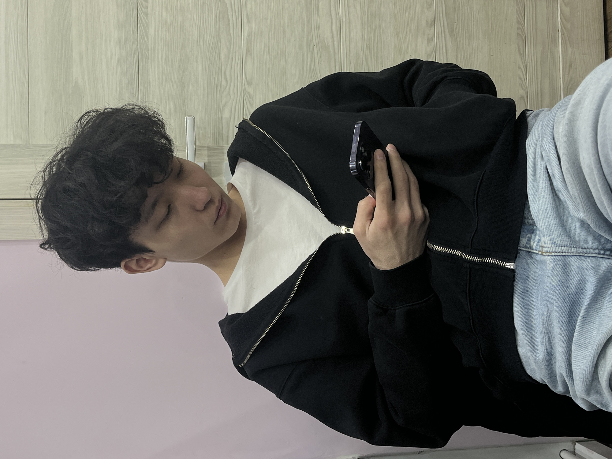 A person with dark, curly hair is holding a smartphone, looking at the screen. The person is wearing a black zip-up hoodie, a white t-shirt underneath, and light blue jeans. The background consists of a light purple wall and a beige wooden door. The location appears to be indoors.