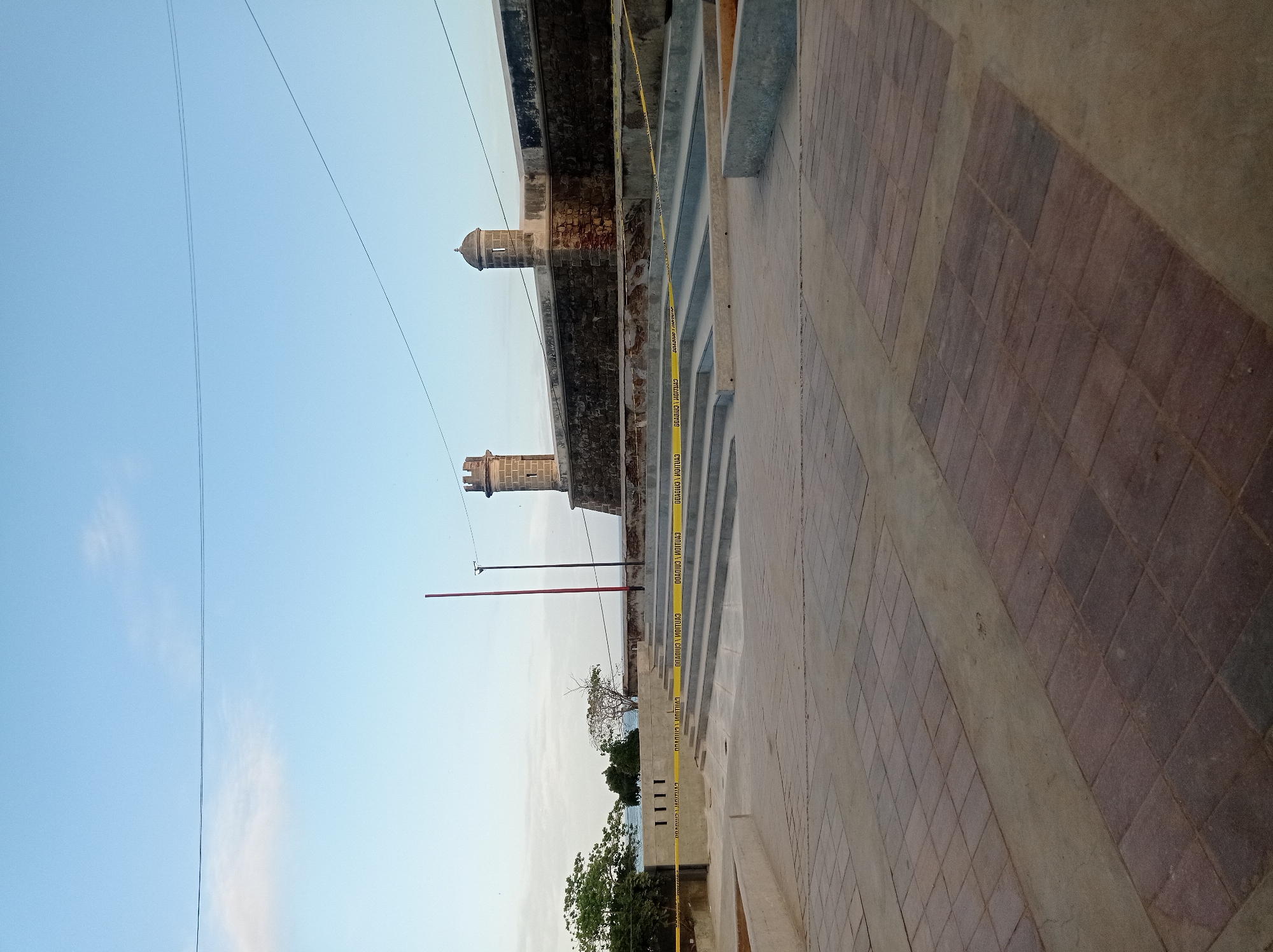 The image shows a partial view of the Castillo San Carlos Borromeo in Pampatar, Venezuela. Two of the fort's turrets are visible against a clear, light blue sky. A stone structure with steps leads to the fort, with yellow caution tape running along the steps. The ground is paved with alternating sections of concrete and reddish-brown tiles. Green trees and vegetation are visible at the base of the fort. Power lines cut across the upper left portion of the sky. A red pole is visible behind the trees.