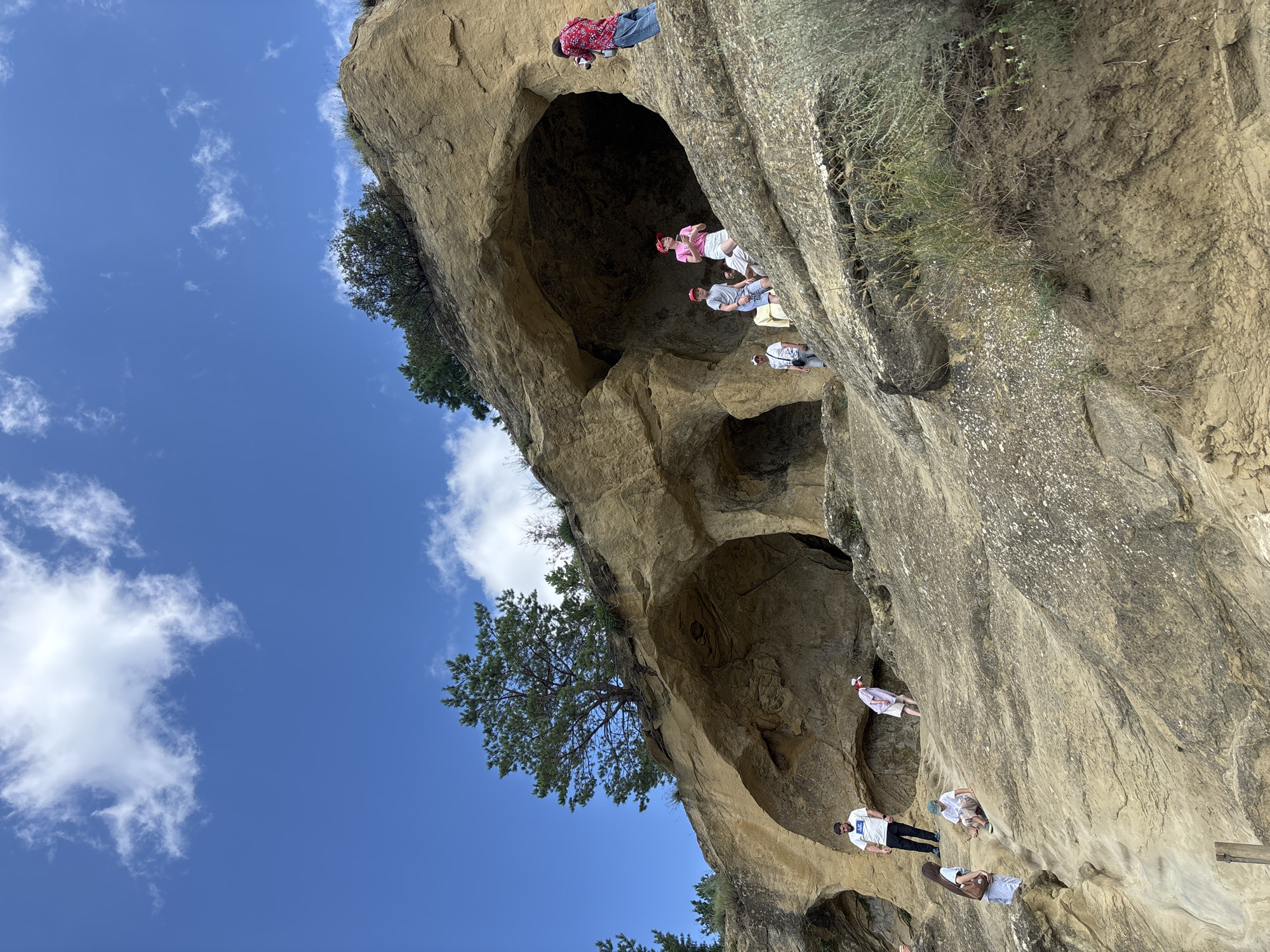 The image depicts a group of people exploring a natural rock formation with several cave-like openings, under a bright blue sky with scattered white clouds. The rock face is a light tan color and appears to be sandstone, with textured surfaces and some vegetation growing on it.
There are approximately eight individuals visible, scattered across the rock face. Some are standing, some are walking, and one person on the far right is holding a camera, seemingly taking a photograph of the cave openings. The people are dressed in casual clothing, suggesting a recreational activity like hiking or sightseeing.
The rock formation itself is the primary subject of the image, with its natural contours and openings dominating the scene. The environment appears to be outdoors, with the natural landscape being the focus. The presence of people suggests it is a location accessible to visitors. The geological formations and the overall setting evoke a sense of natural exploration and history.