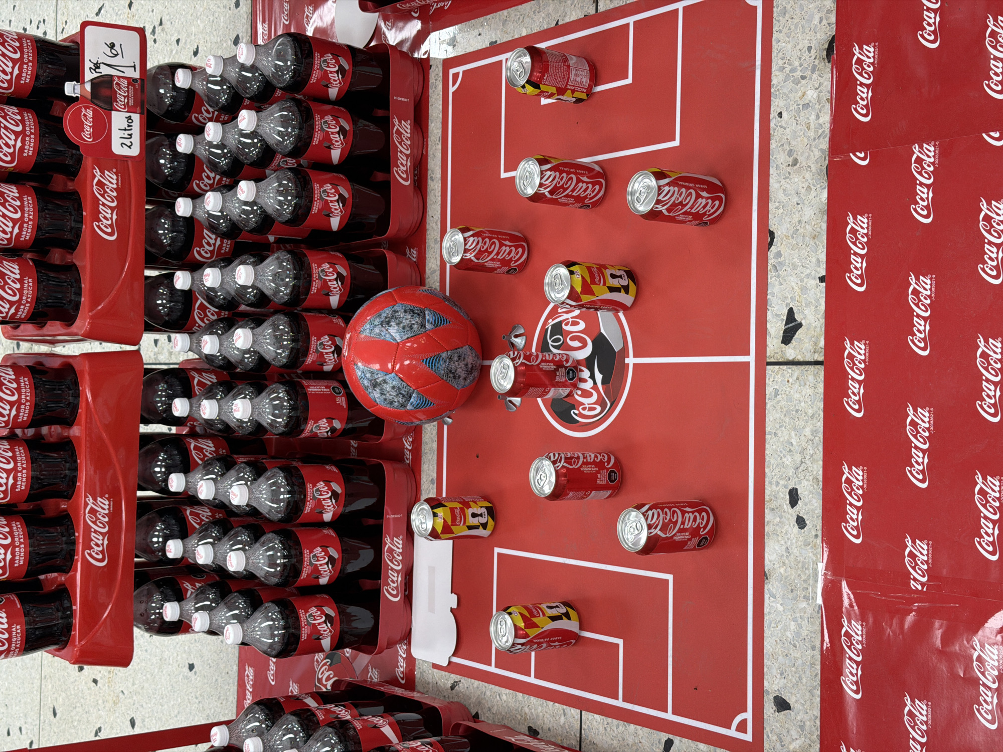 The image depicts a promotional display for Coca-Cola products, set up in what appears to be a retail environment. The display features several shelves stacked with Coca-Cola bottles and cans. In the foreground, a red surface designed to resemble a soccer field is laid out, with white lines marking boundaries and a penalty box. Several Coca-Cola cans are strategically placed on this "field" as if representing players. A soccer ball, also red and blue, is positioned on the left side of the field. The background shows more Coca-Cola products on tiered shelving, with some bottles having red caps and others white. The overall theme combines Coca-Cola branding with a soccer motif, possibly for a promotional event. The location is identified as Turmero, Venezuela. The scene is static, with no people or overt actions depicted. The lighting suggests an indoor commercial setting.