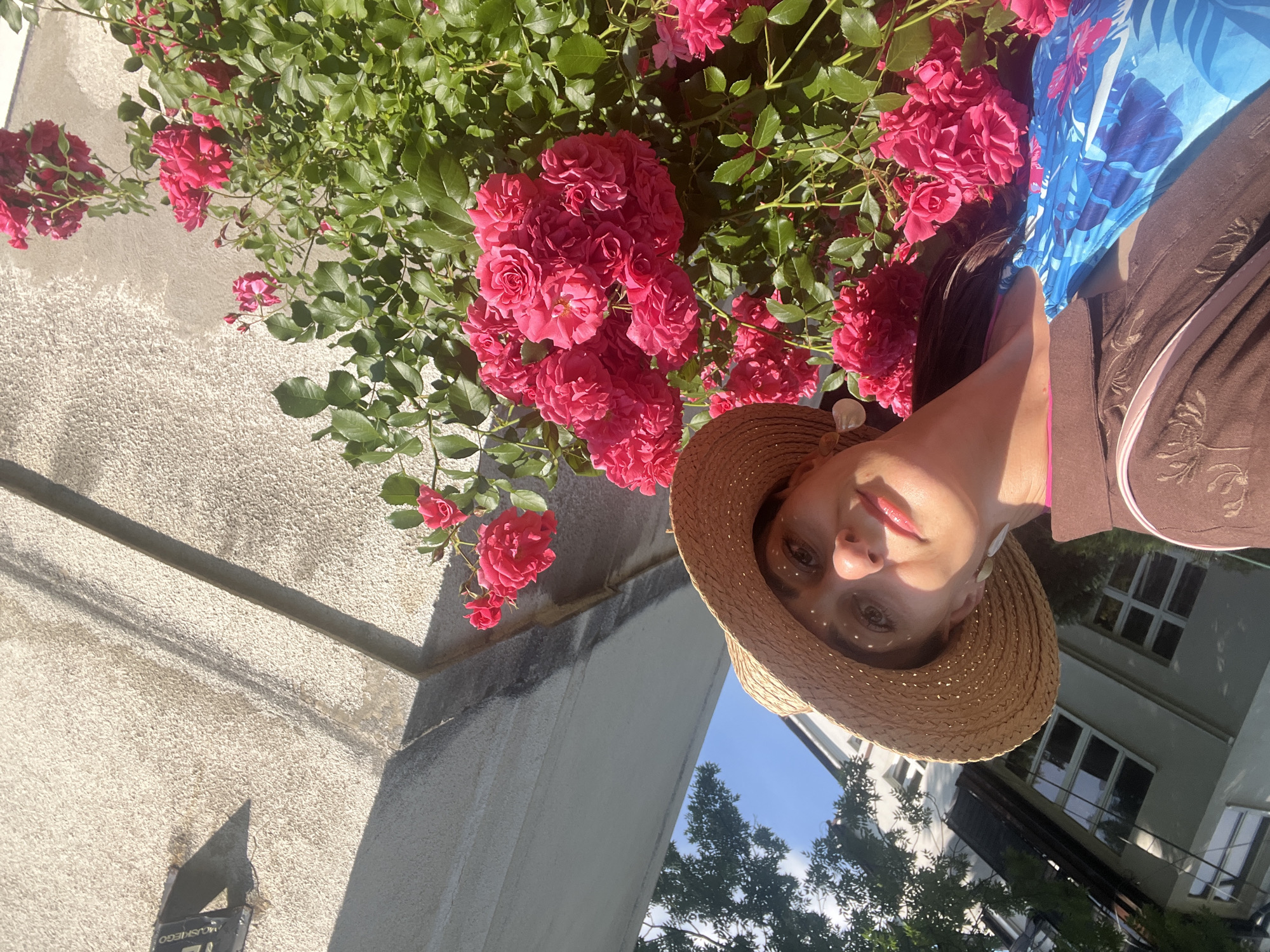 In an outdoor urban setting, a woman is captured in a selfie next to a bush of bright pink roses. She wears a straw hat, a brown patterned top, and a blue and white floral garment. Her earrings are visible, one of which is a white shell-shaped dangle. The background features a textured concrete wall with a plaque at the top, and a building with windows is partially visible to the left, with green foliage from a tree overhead. The scene appears to be under a clear blue sky on a sunny day.